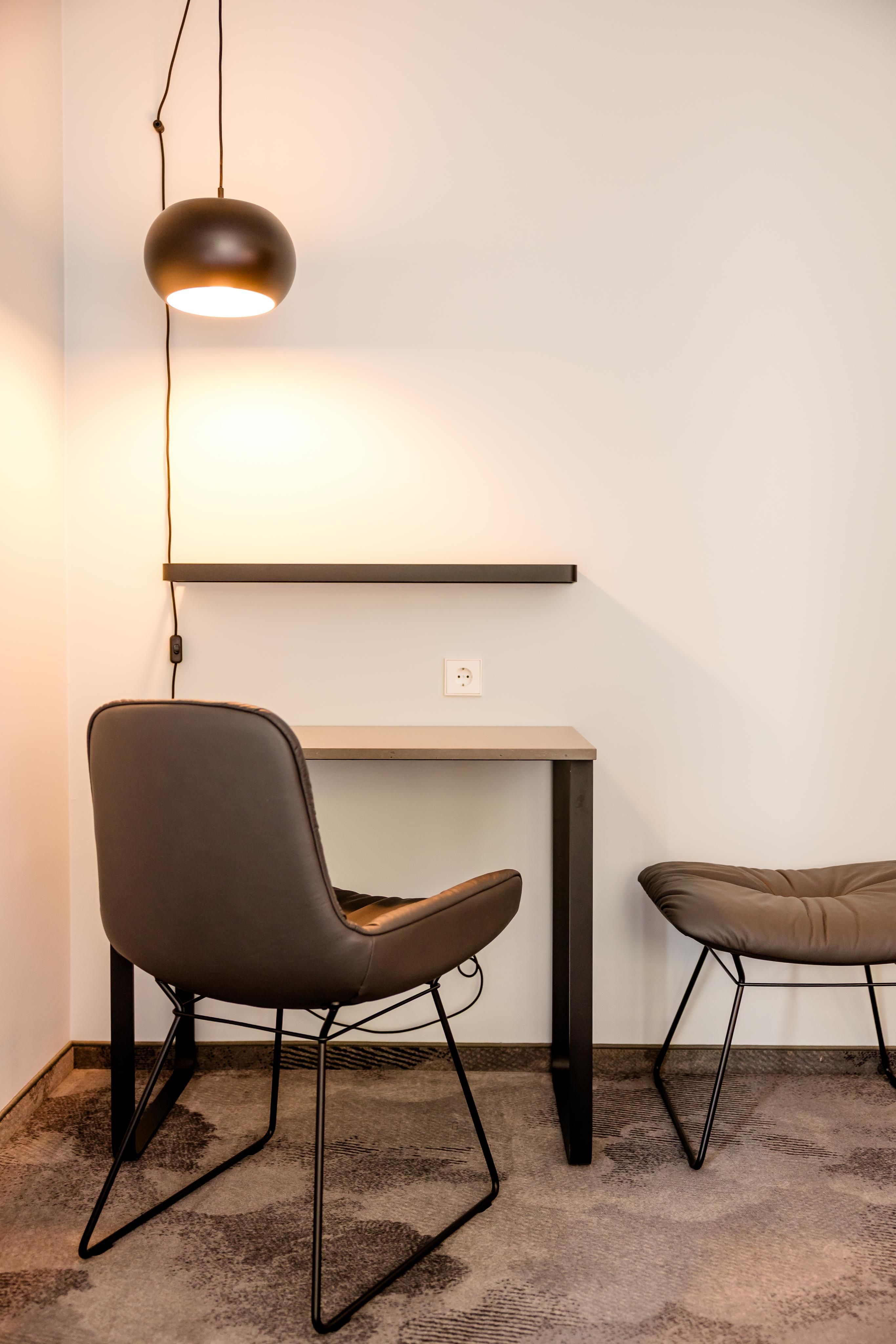 Moderner Arbeitsplatz mit kleinem Schreibtisch, bequemen Stuhl und Hocker, dazu eine warme Hängelampe für angenehmes Licht, ideal zum konzentrierten Arbeiten oder Lesen im ruhigen Raum.