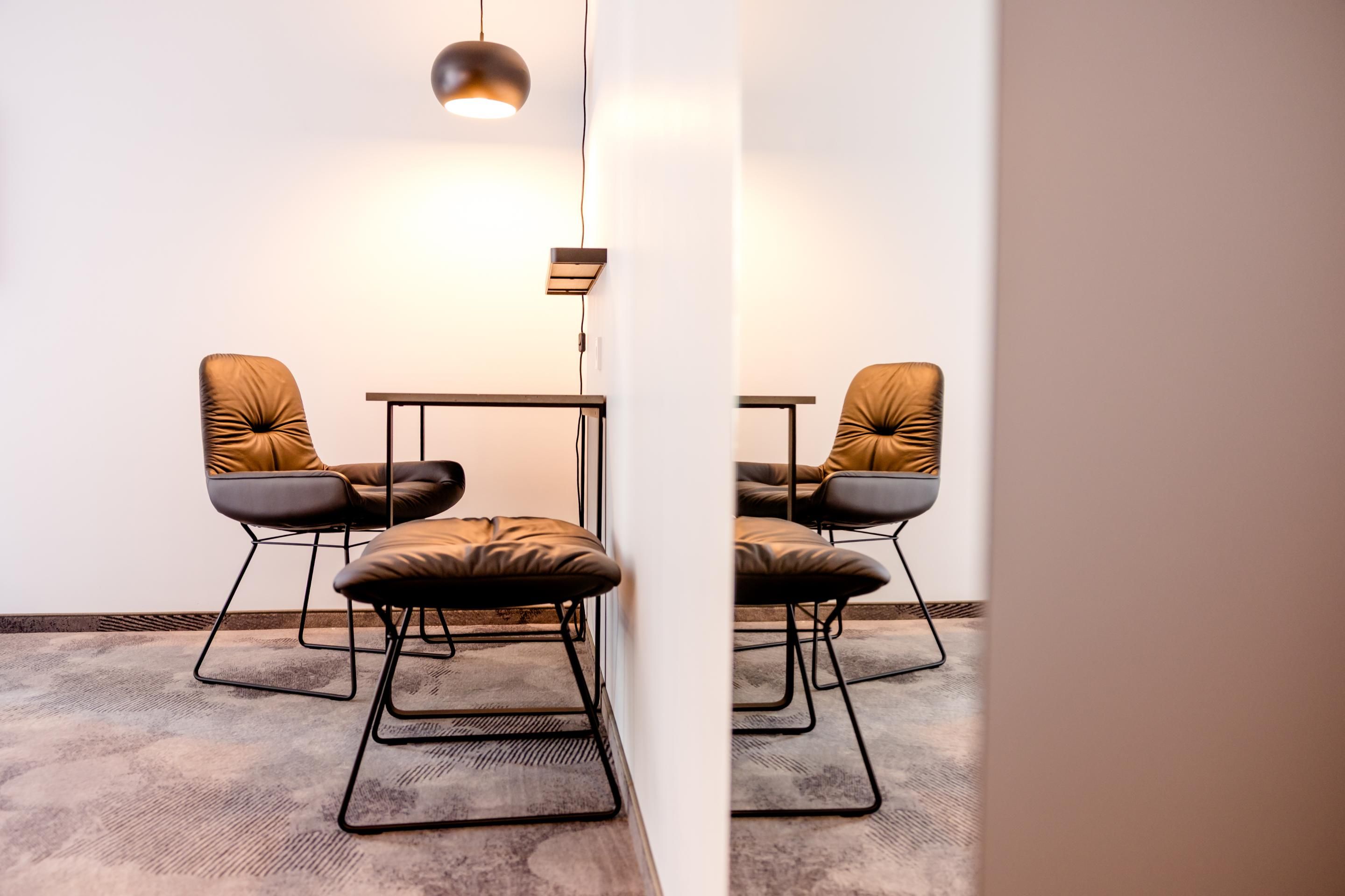 Moderner, gemütlicher Sitzbereich mit zwei gepolsterten Lederstühlen, einem passenden Hocker und einem kleinen Tisch, sanft beleuchtet von einer hängenden Lampe vor einer hellen Wand, die durch einen Spiegel reflektiert wird.