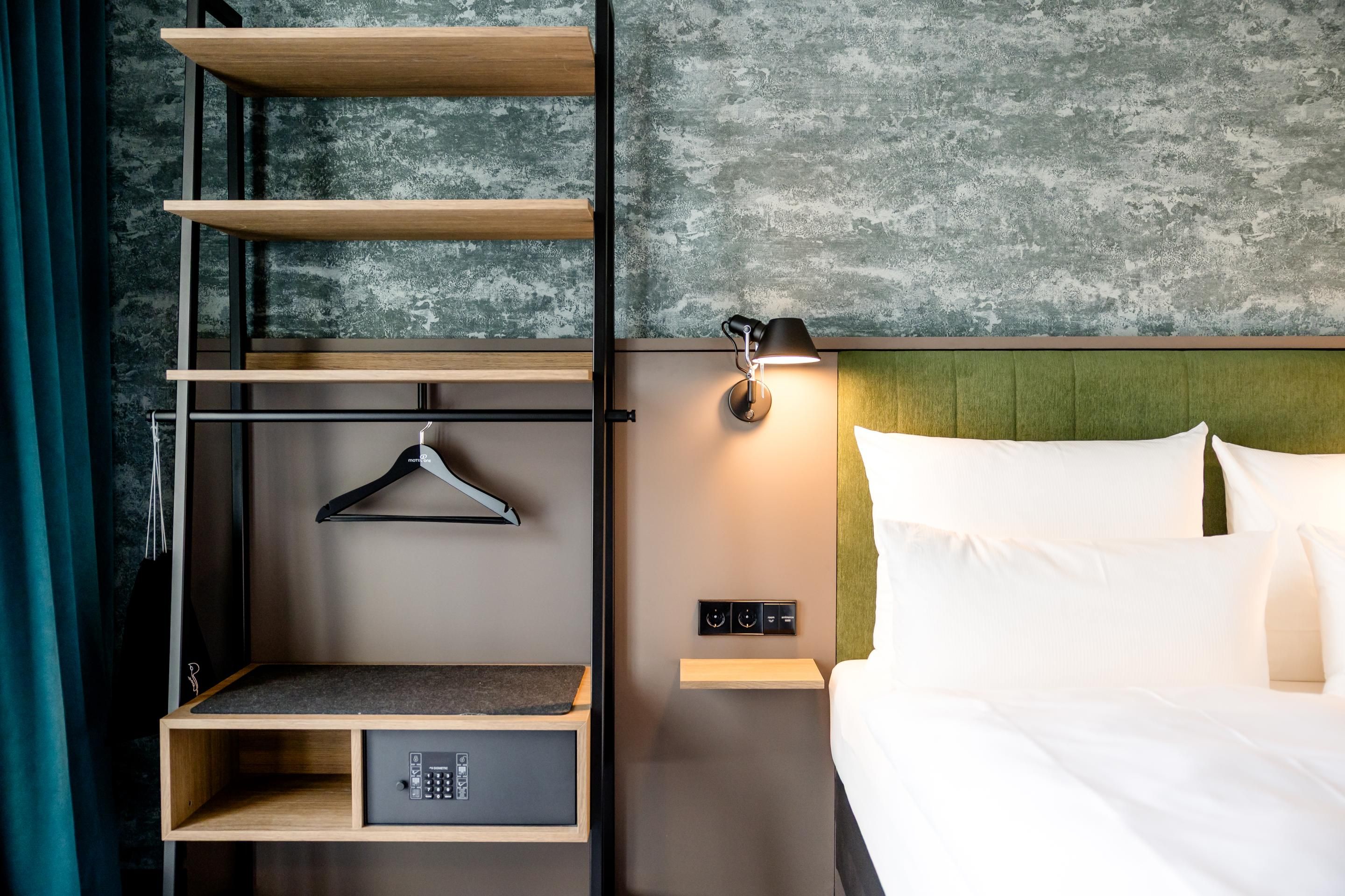 Moderner Hotelzimmerbereich mit grün gepolstertem Bettkopfteil, weißer Bettwäsche, offener Garderobe mit Kleiderbügeln und integriertem Safe, dezenter Wandlampe und stilvoller, minimalistischer Einrichtung.