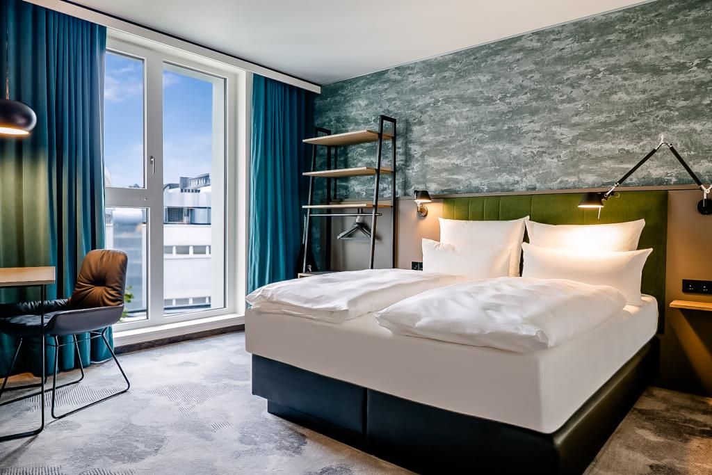 Modernes Hotelzimmer mit großem Doppelbett, sanfter Beleuchtung und urbanem Ausblick. Ruhige Atmosphäre, stilvolle Einrichtung mit grünem Akzent, ideal für erholsamen Aufenthalt. Große Fenster lassen viel Tageslicht herein, komfortabler Arbeitsplatz mit Stuhl am Fenster.
