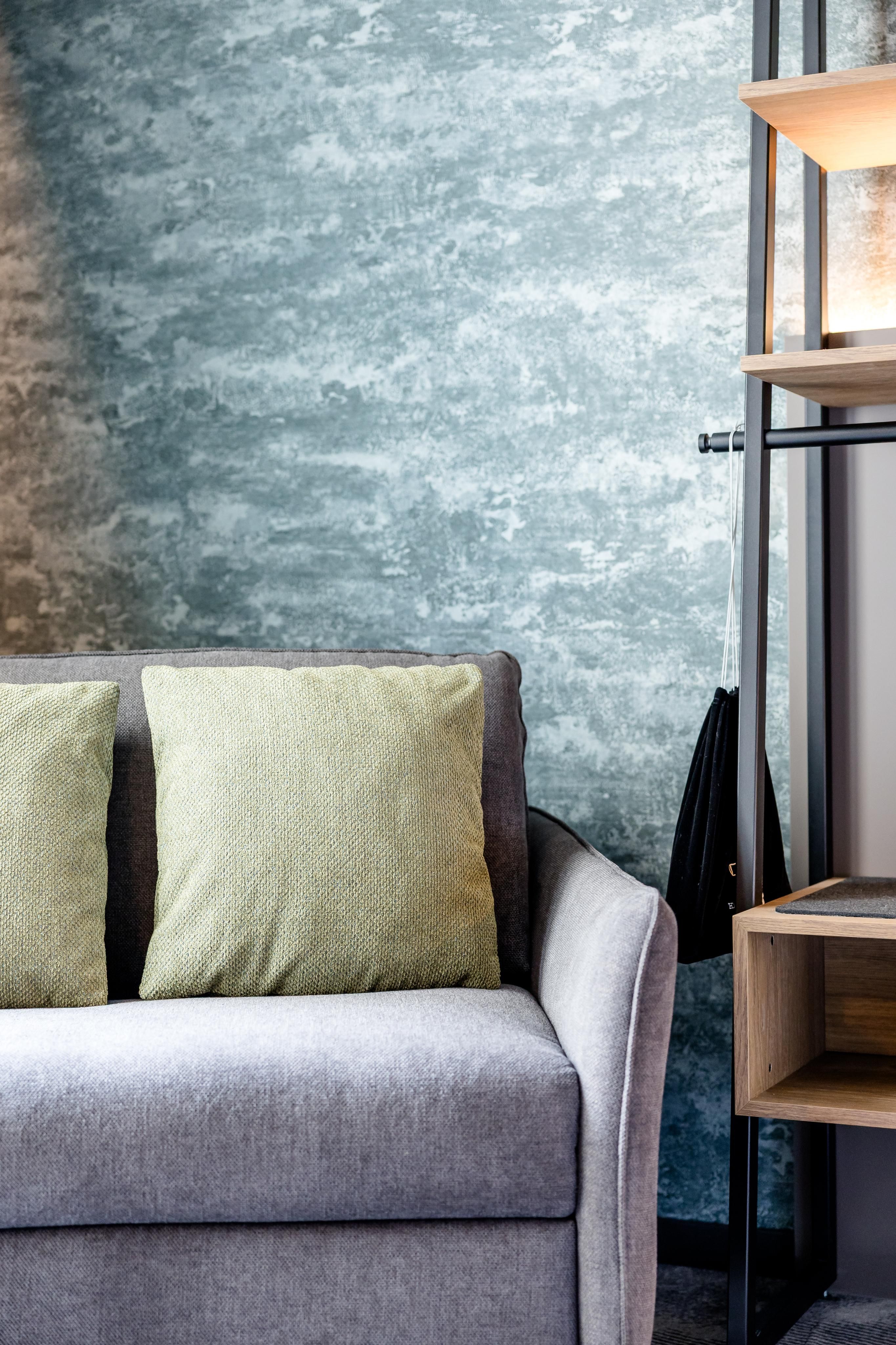 Gemütliche Sitzecke mit grauem Sofa und hellgrünen Kissen vor einer strukturierten, blaugrauen Wand, daneben modernes Holzregal mit indirekter Beleuchtung, ideal für entspannte Momente im Zimmer.