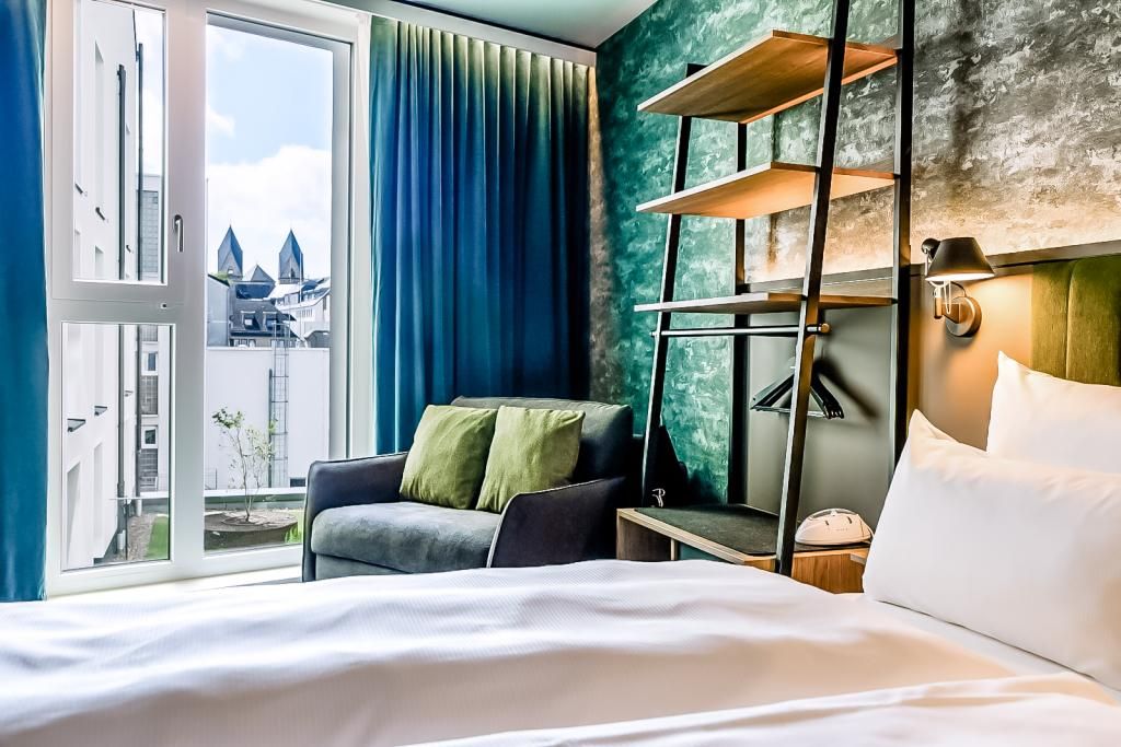Modernes Hotelzimmer mit gemütlichem Bett, grünem Akzentwanddesign, bequemen Sesseln und großem Fenster, das Tageslicht und Aussicht auf Stadtgebäude bietet.