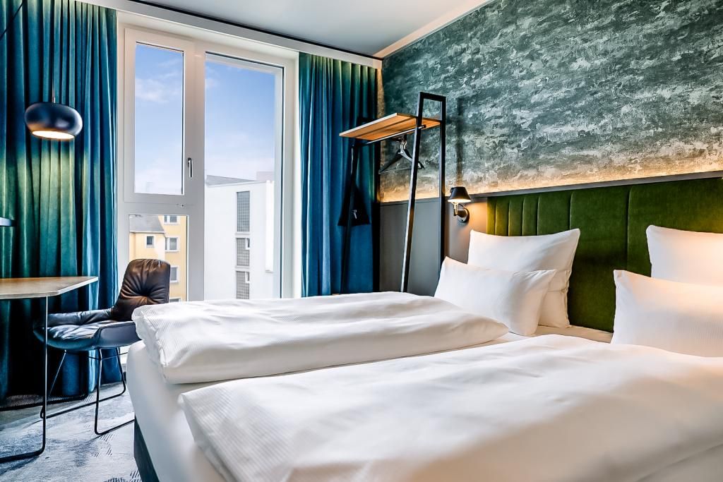 Modernes Hotelzimmer mit zwei Einzelbetten, grünem Akzentkopfteil, großen Fenstern und samtigen Vorhängen. Helle, freundliche Atmosphäre mit stilvollem Design und komfortabler Ausstattung.