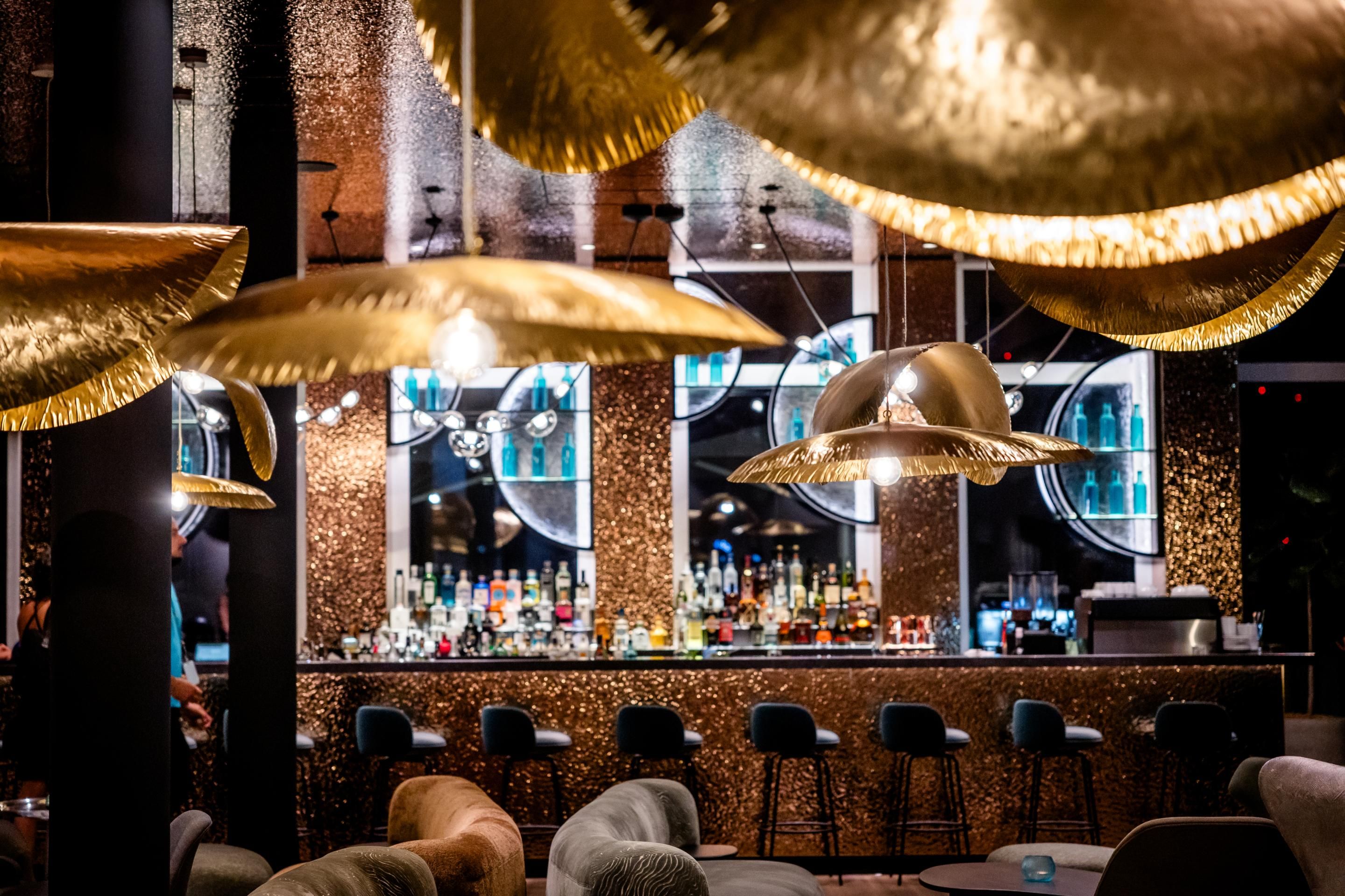 Moderne Hotelbar mit elegantem, golden schimmerndem Interieur, stilvollen Barhockern und gemütlichen Sitzbereichen, die eine luxuriöse und einladende Atmosphäre für entspannte Abende bieten.