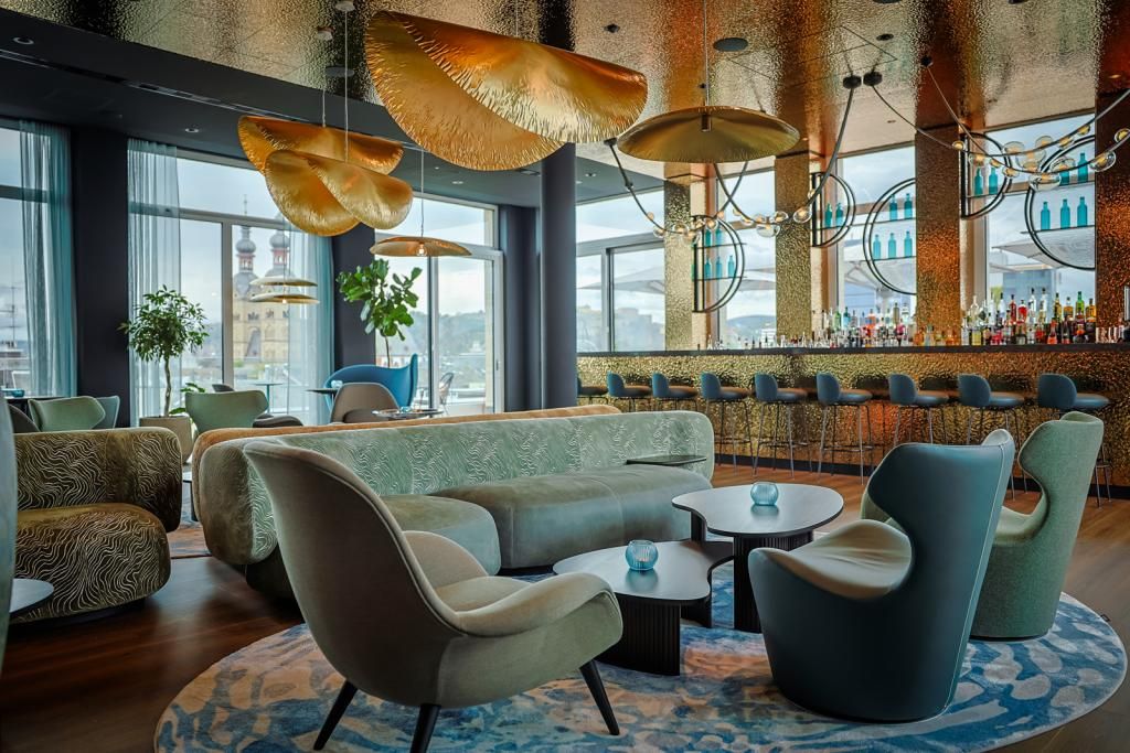 Elegante Hotelbar mit bequemen Sesseln und Sofas, modernen goldfarbenen Deckenleuchten, Panoramafenstern mit Stadtblick und stilvoller Atmosphäre für entspannte Abende und geselliges Beisammensein.