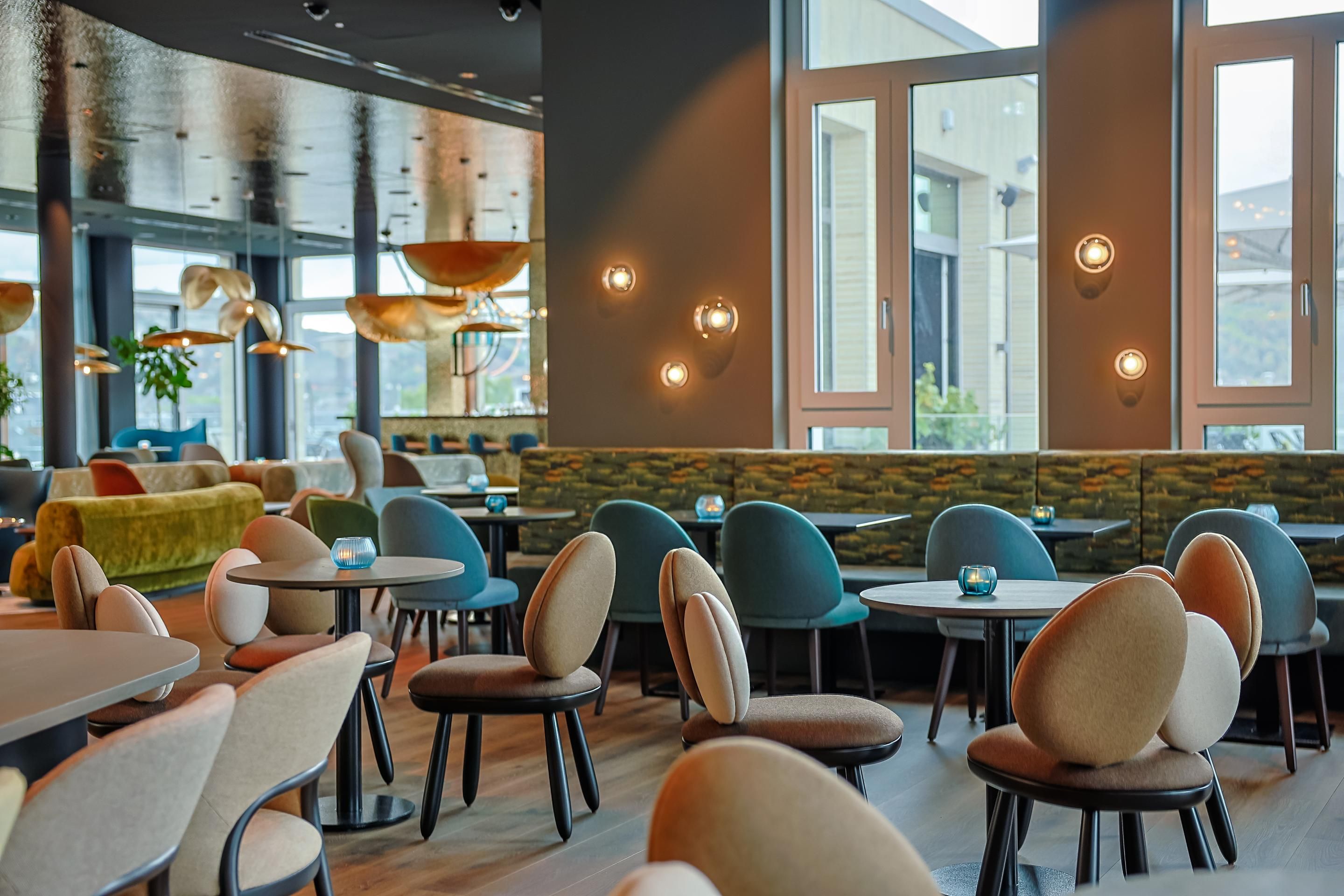 Modern eingerichteter Hotelrestaurantbereich mit stilvollen Stühlen, runden Tischen und großen Fenstern, die Tageslicht hereinlassen. Atmosphärische Wandbeleuchtung und dezente Dekoration schaffen eine einladende, gemütliche Stimmung für Gäste.