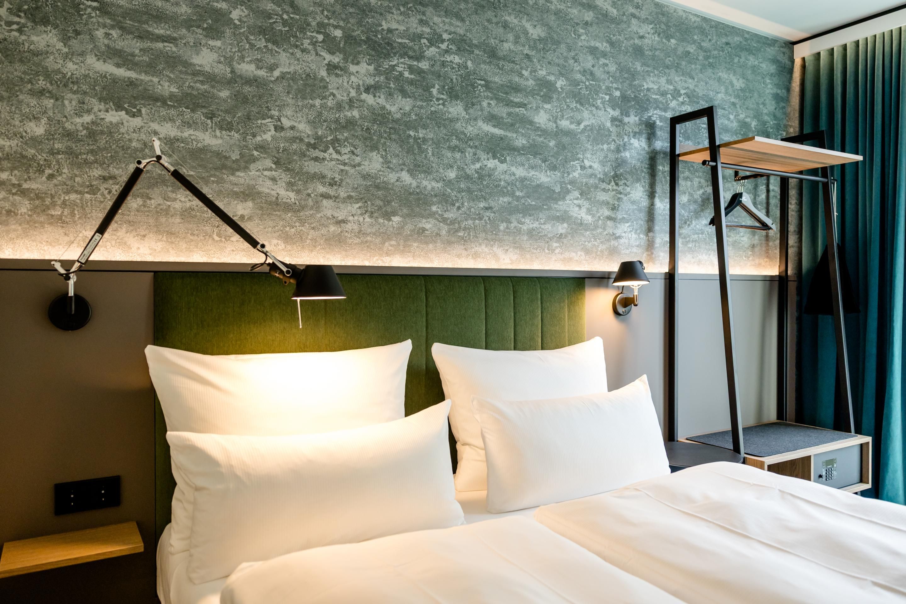 Modern eingerichtetes Hotelzimmer mit gemütlichem Doppelbett, grünem gepolstertem Kopfteil, warmem Licht und offenem Garderobenständer für bequemen Stauraum.