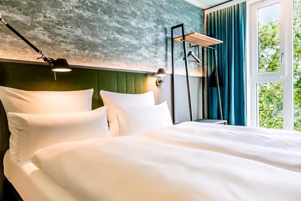 Helles, modern eingerichtetes Hotelzimmer mit großem, komfortablem Doppelbett, grüner Akzentwand, indirekter Beleuchtung und großem Fenster mit Blick ins Grüne.