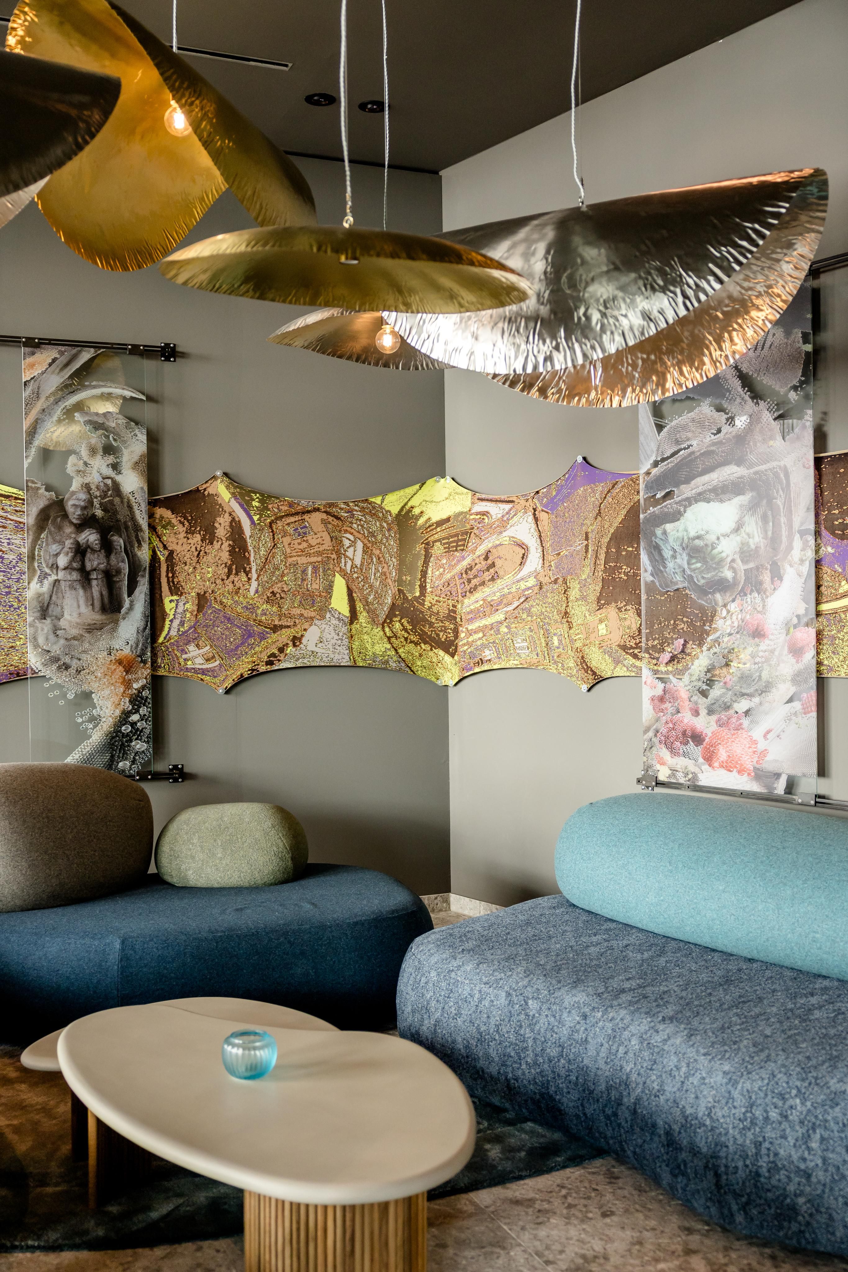 Moderner Lounge-Bereich mit geschwungenen Sofas in Blau- und Grautönen, ovalem Couchtisch und dekorativen, goldfarbenen Hängelampen, ergänzt durch kunstvolle Wanddekorationen mit abstrakten Motiven.