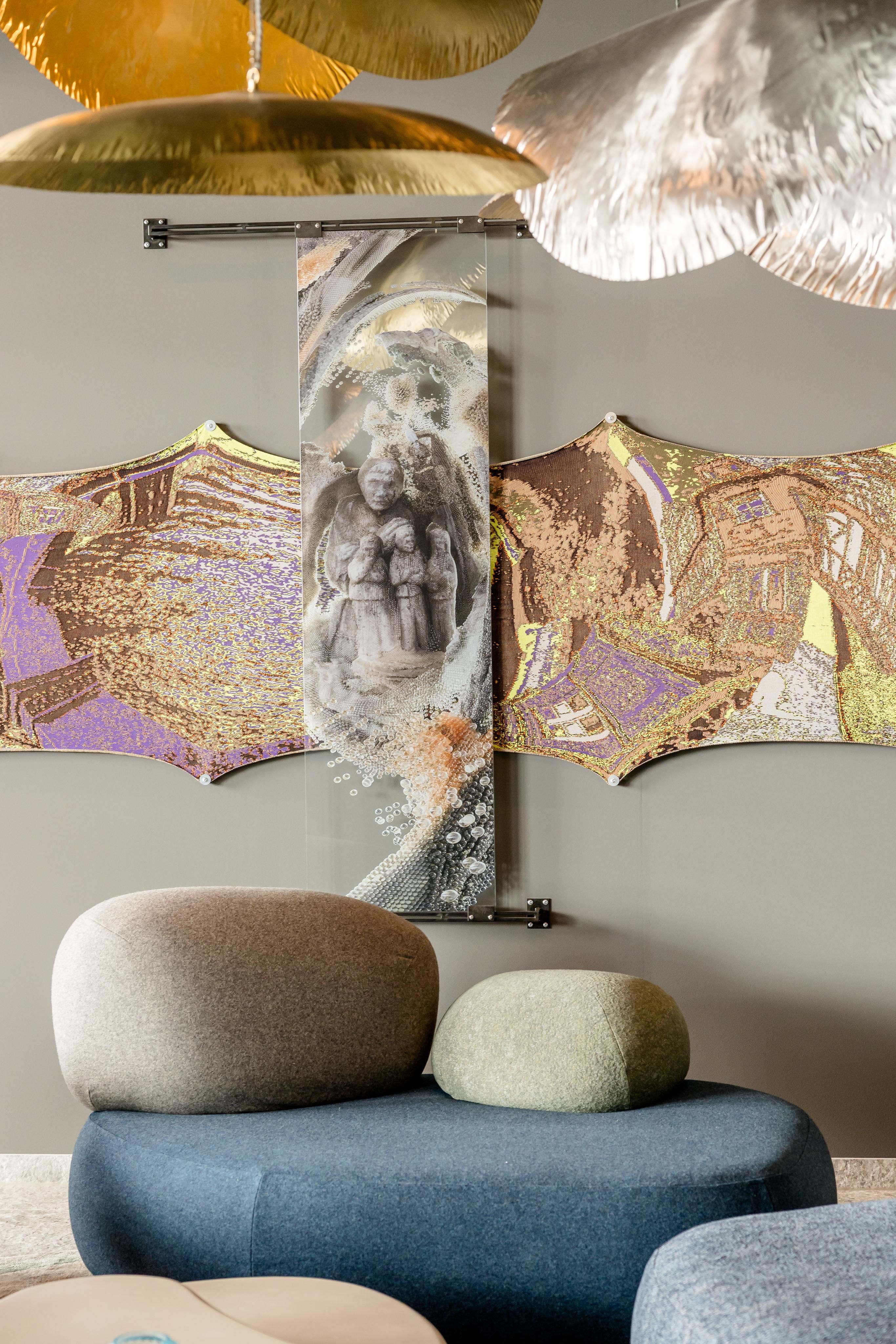 Moderner Sitzbereich mit abgerundeten, gepolsterten Sitzmöbeln in sanften Grau- und Blautönen. Im Hintergrund abstrakte Wandkunst mit goldenen und violetten Akzenten. Decke mit großen, metallisch schimmernden, dekorativen Elementen schafft ein elegantes Ambiente.