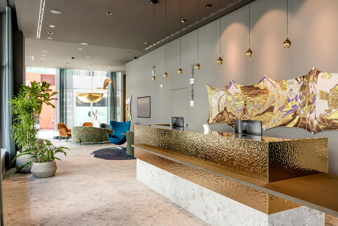 Moderner Hotel-Empfang mit goldglänzendem Empfangstresen, stilvollen Pendelleuchten und Pflanzen. Im Hintergrund gemütliche Sitzbereiche mit großen Fenstern für viel Tageslicht. Elegante, einladende Atmosphäre für ankommende Gäste.