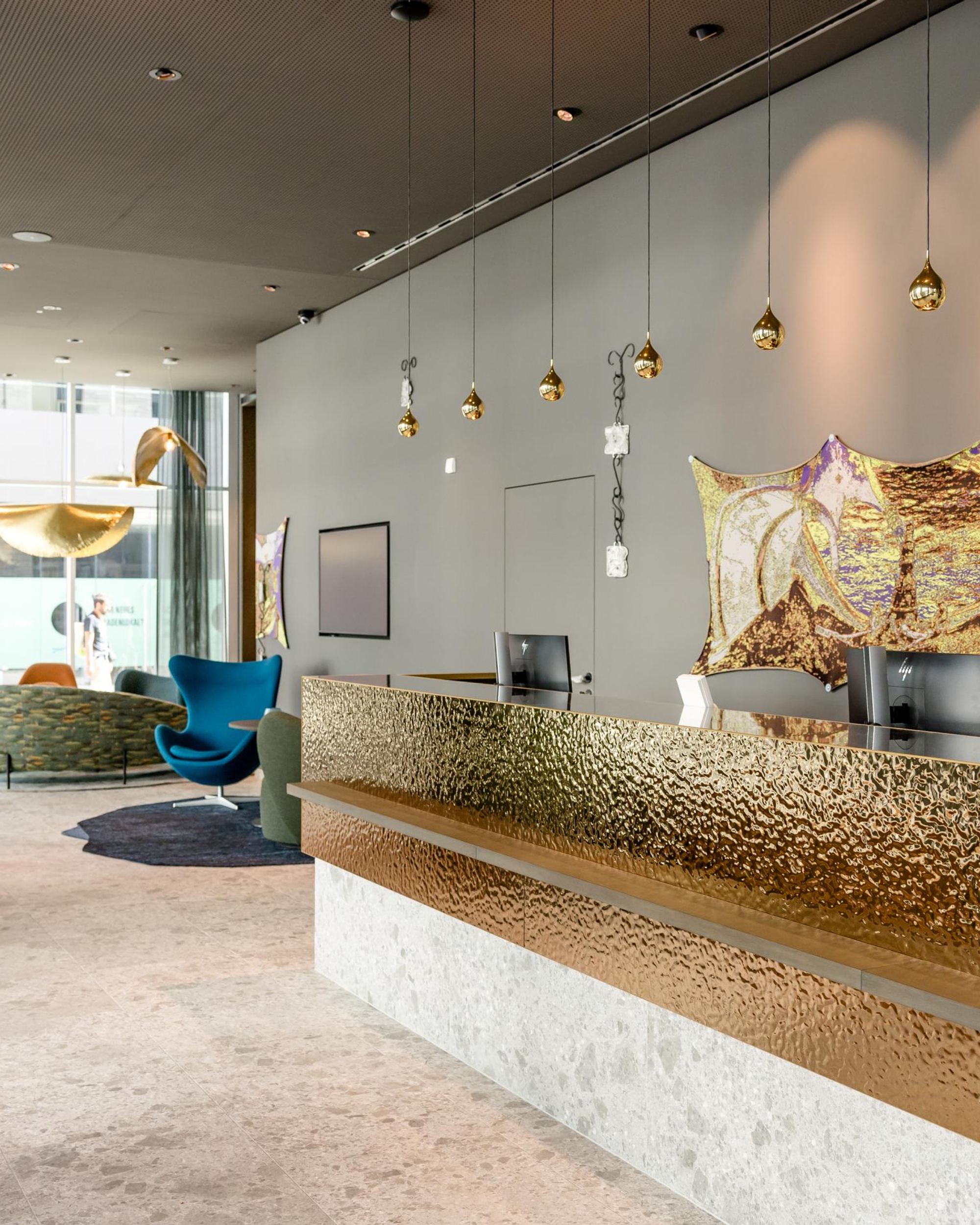 Moderner Hotel-Empfang mit goldglänzendem Empfangstresen, stilvollen Pendelleuchten und Pflanzen. Im Hintergrund gemütliche Sitzbereiche mit großen Fenstern für viel Tageslicht. Elegante, einladende Atmosphäre für ankommende Gäste.