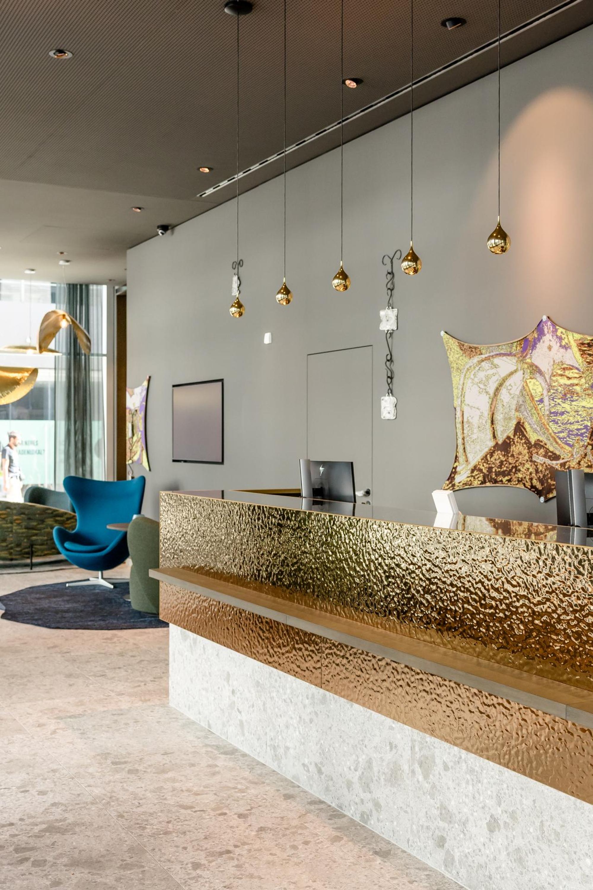 Moderner Hotel-Empfang mit goldglänzendem Empfangstresen, stilvollen Pendelleuchten und Pflanzen. Im Hintergrund gemütliche Sitzbereiche mit großen Fenstern für viel Tageslicht. Elegante, einladende Atmosphäre für ankommende Gäste.