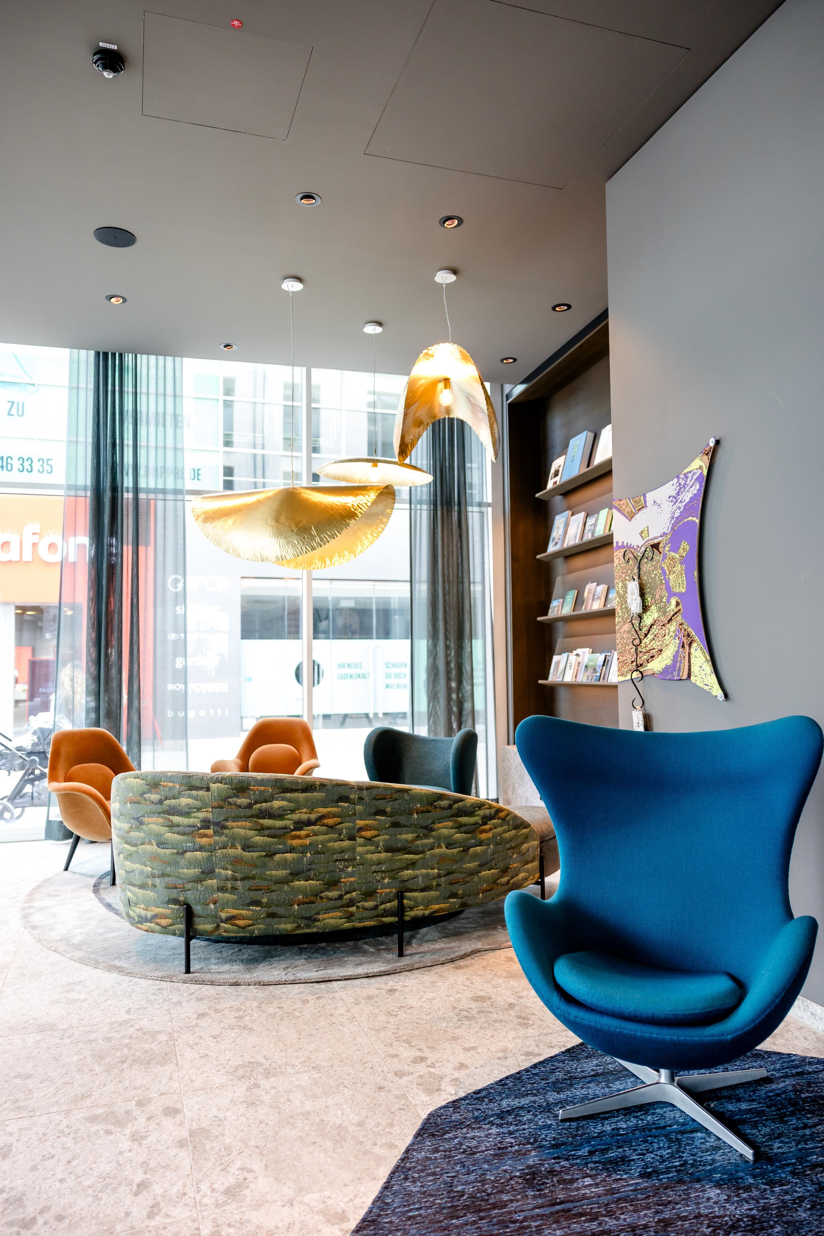 Moderner, heller Lounge-Bereich mit stylischen Sitzmöbeln, darunter ein blauer Design-Sessel und ein geschwungene grüne Sofainsel. Große Fenster lassen natürliches Licht herein, goldfarbene Pendelleuchten hängen von der Decke, eine Wand mit Büchern und Kunst ergänzt die entspannte Atmosphäre.