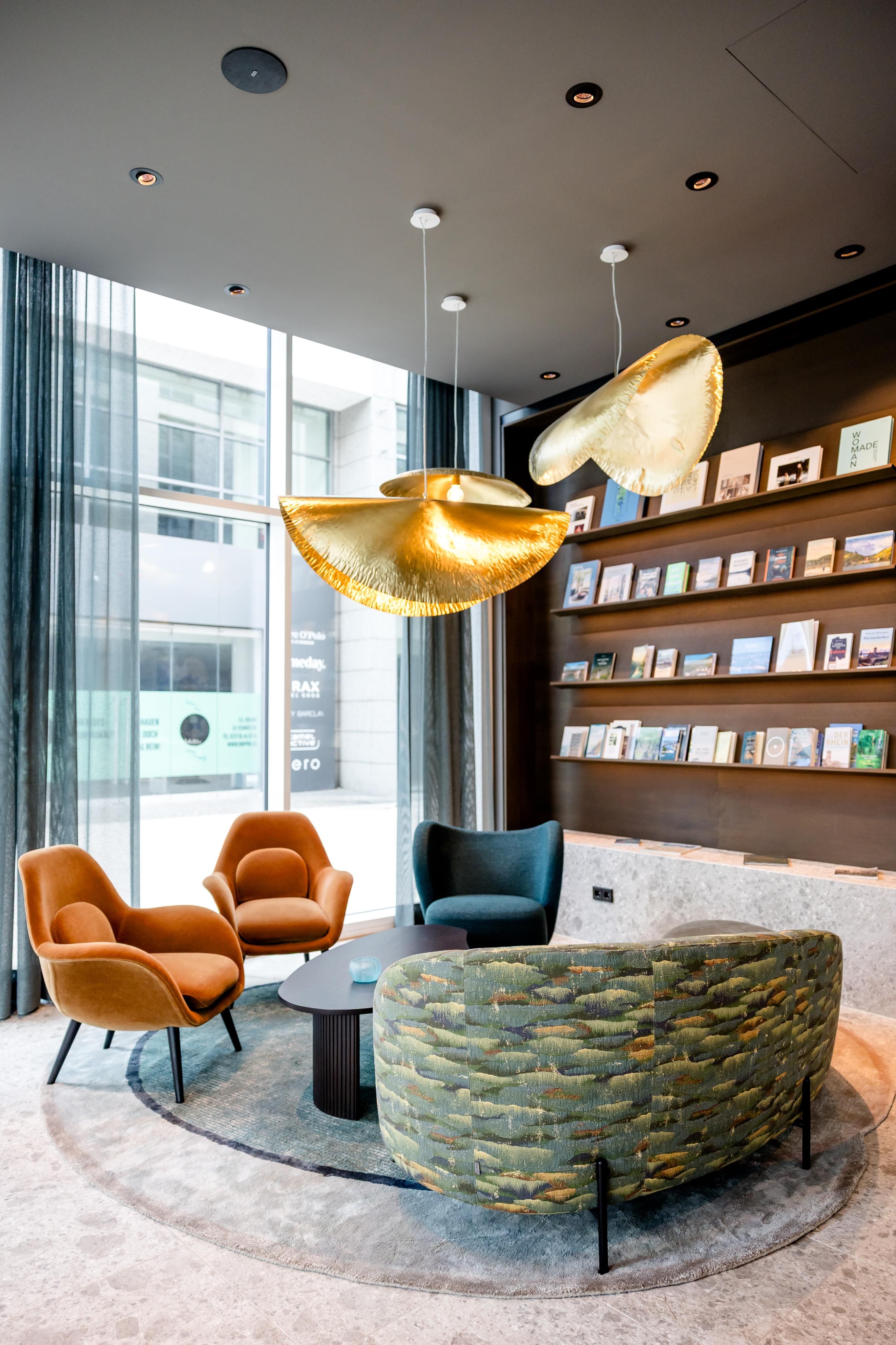 Moderner Hotel-Lounge-Bereich mit großen Fenstern, gemütlichen Sesseln in Orange und Blau, gerundeter Couch mit Muster, runden Teppich und auffälligen goldfarbenen Hängeleuchten für entspanntes Wohlfühlambiente.
