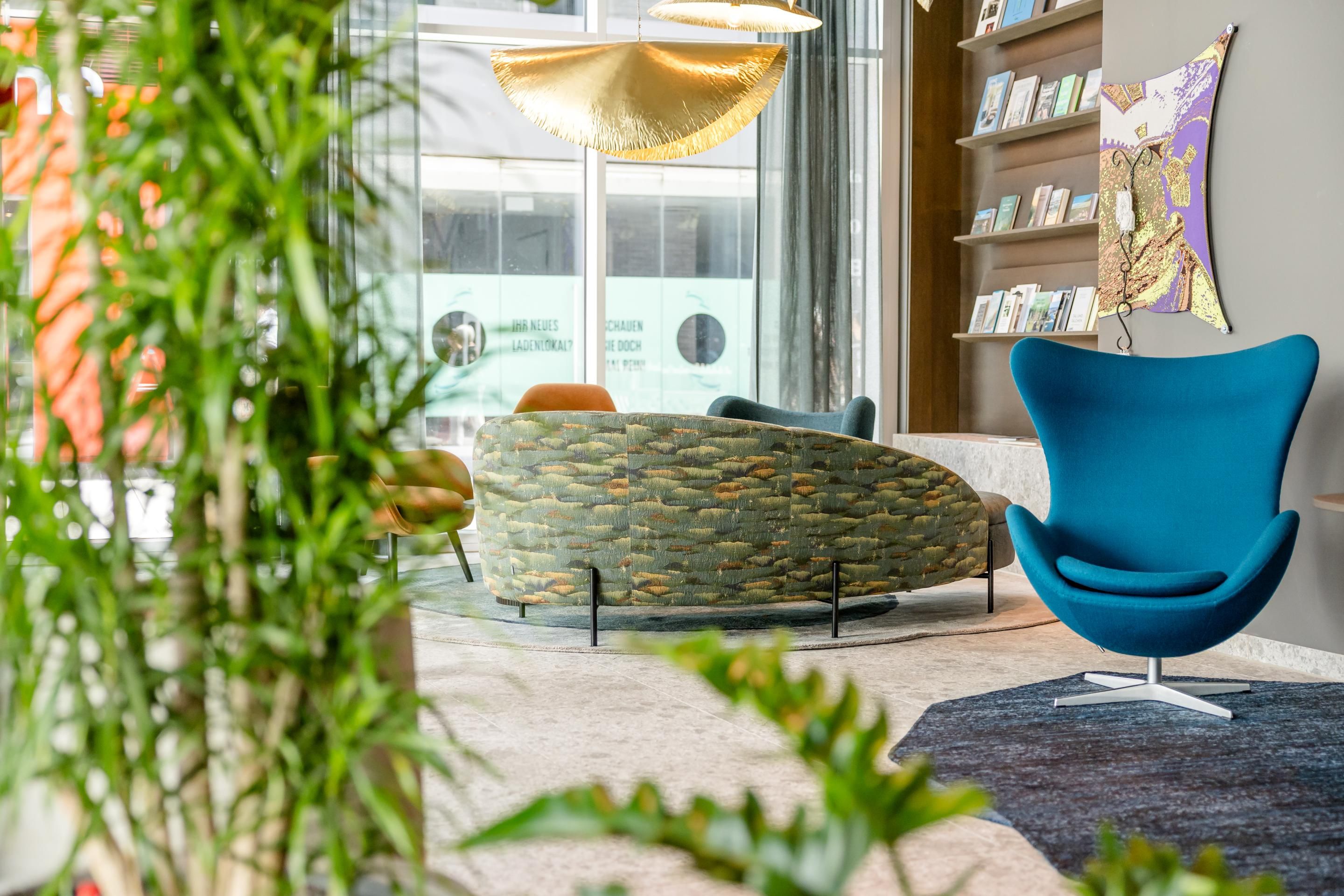 Moderner Lounge-Bereich mit bequemem, blauem Design-Sessel, rundem Sofa und kreativem Wanddekor, umgeben von grünen Pflanzen. Helle, freundliche Atmosphäre durch große Fenster mit Tageslicht.