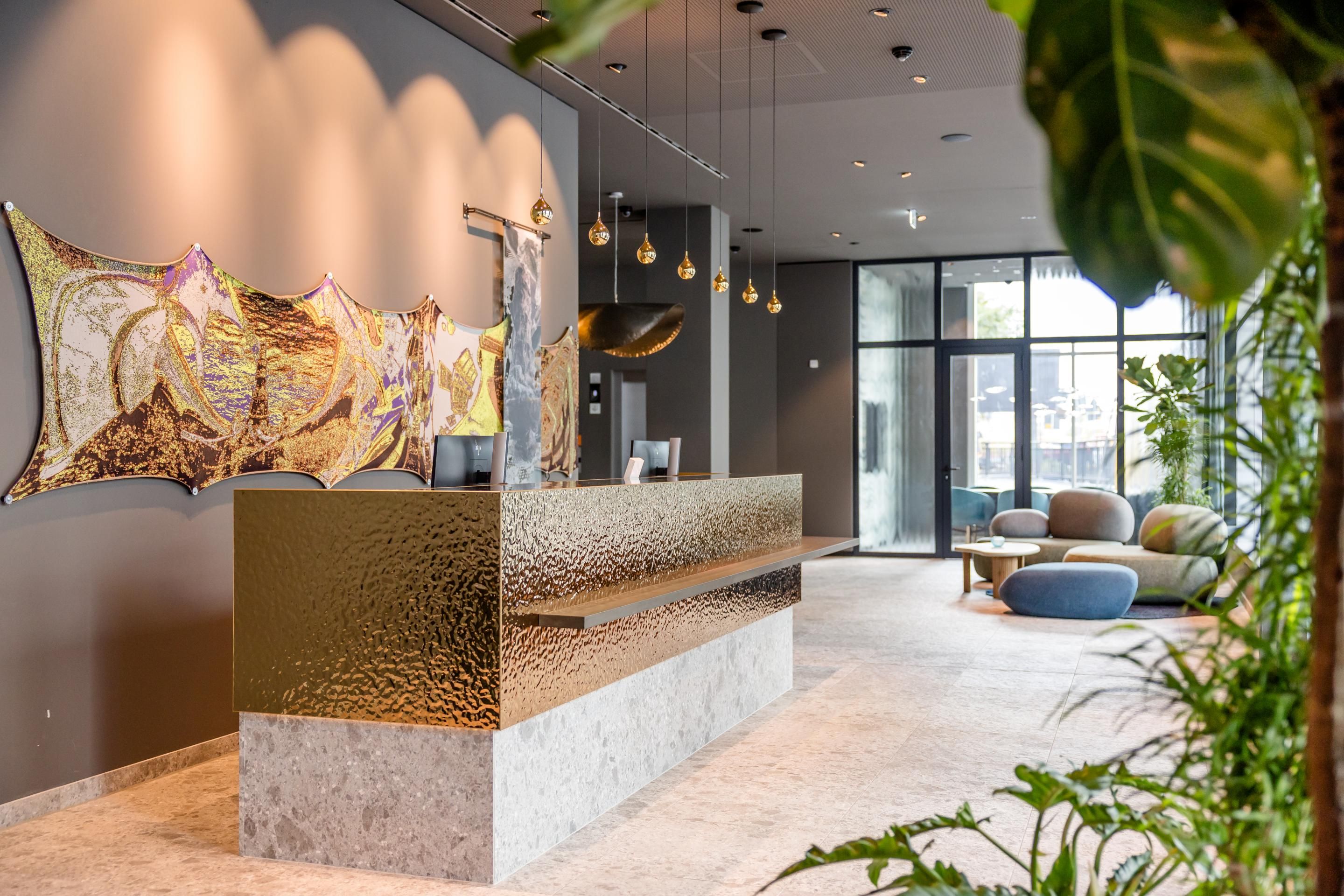 Moderner Hotellobby-Empfang mit goldfarbenem Tresen, warmen Lichtakzenten und stilvoller Wanddekoration, hellem Sitzbereich und grünen Pflanzen, einladend für Gäste.