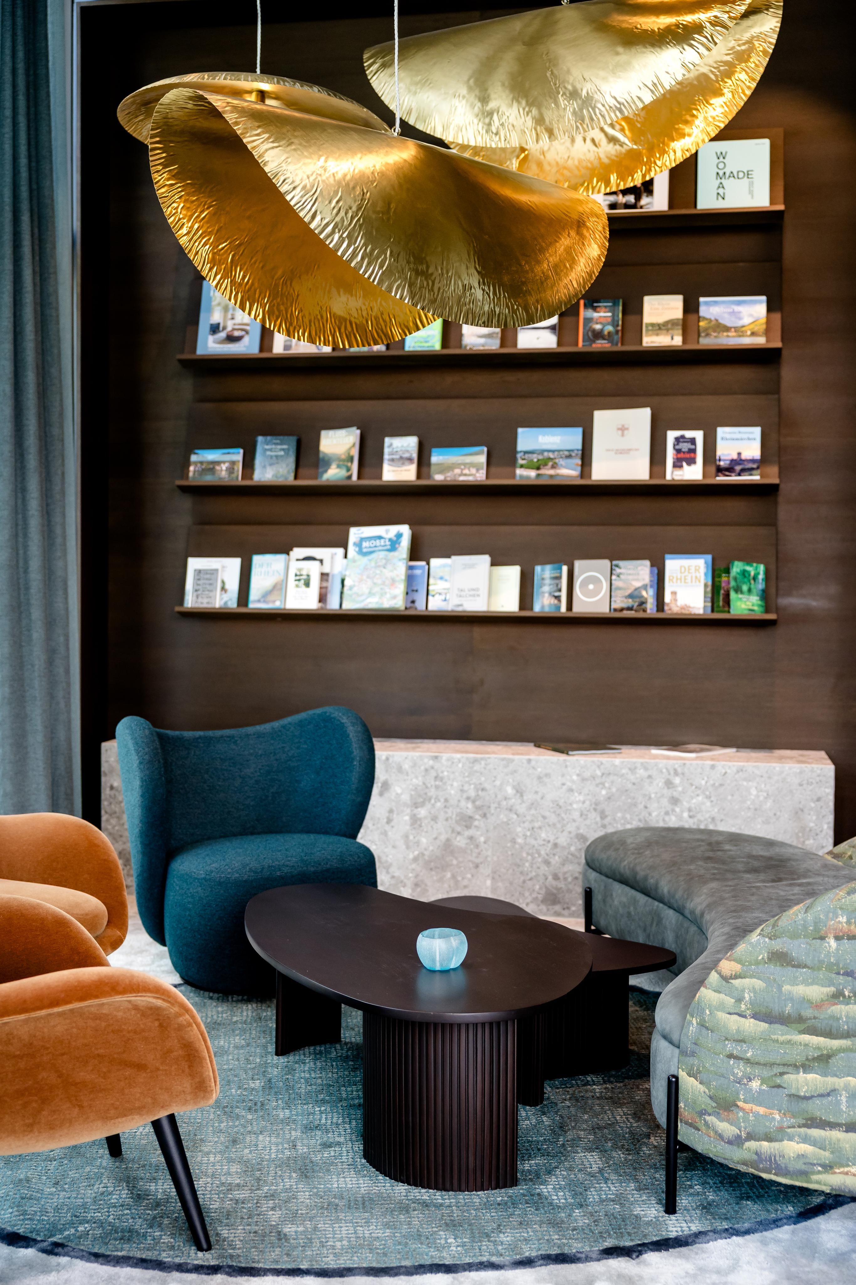Moderne Lounge mit gemütlichen Sesseln und Sofas, elegantem dunklem Holzhintergrund mit Büchern und Broschüren, darunter runder Couchtisch. Zwei große, goldene Designerhängeleuchten schaffen warme Atmosphäre.