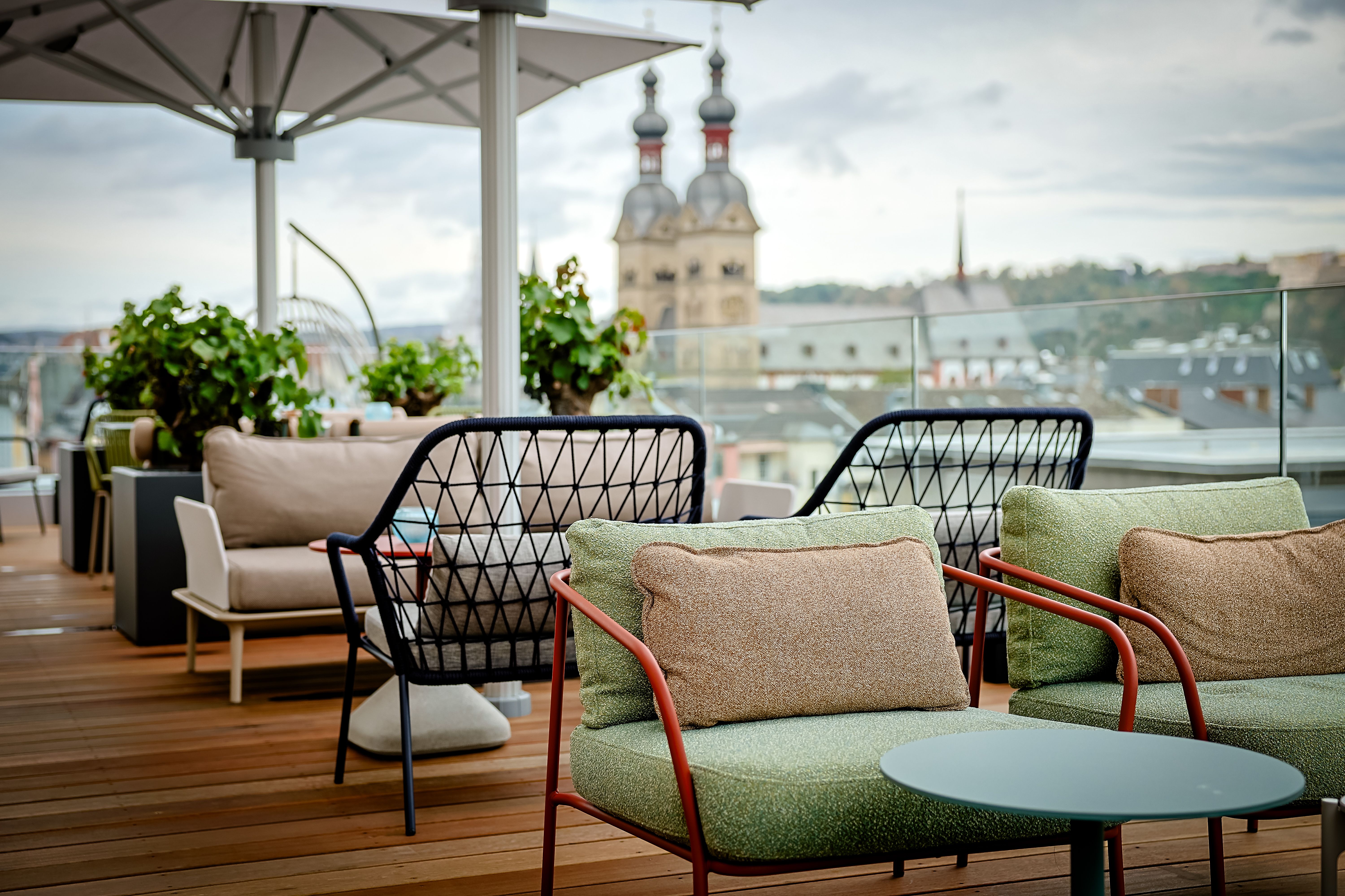 Gemütliche Dachterrassen-Lounge mit gepolsterten Sesseln und Tischen, umgeben von Pflanzen, mit Blick auf historische Kirchtürme in der Stadt. Ideal zum Entspannen und Genießen der Aussicht.
