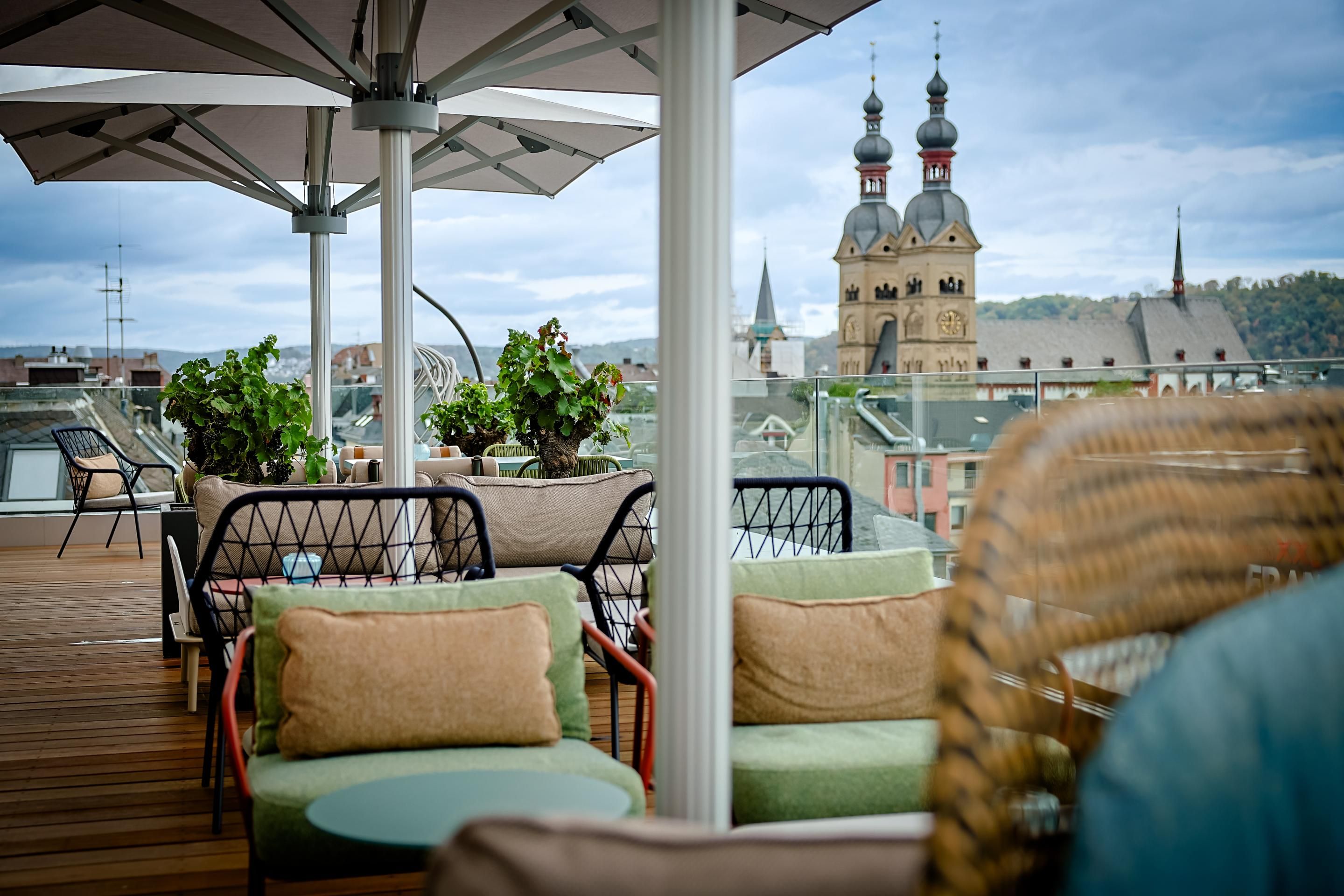 Gemütliche Dachterrasse mit Sitzbereichen und Pflanzen, Blick auf historische Kirchtürme und Stadtpanorama bei bewölktem Himmel.