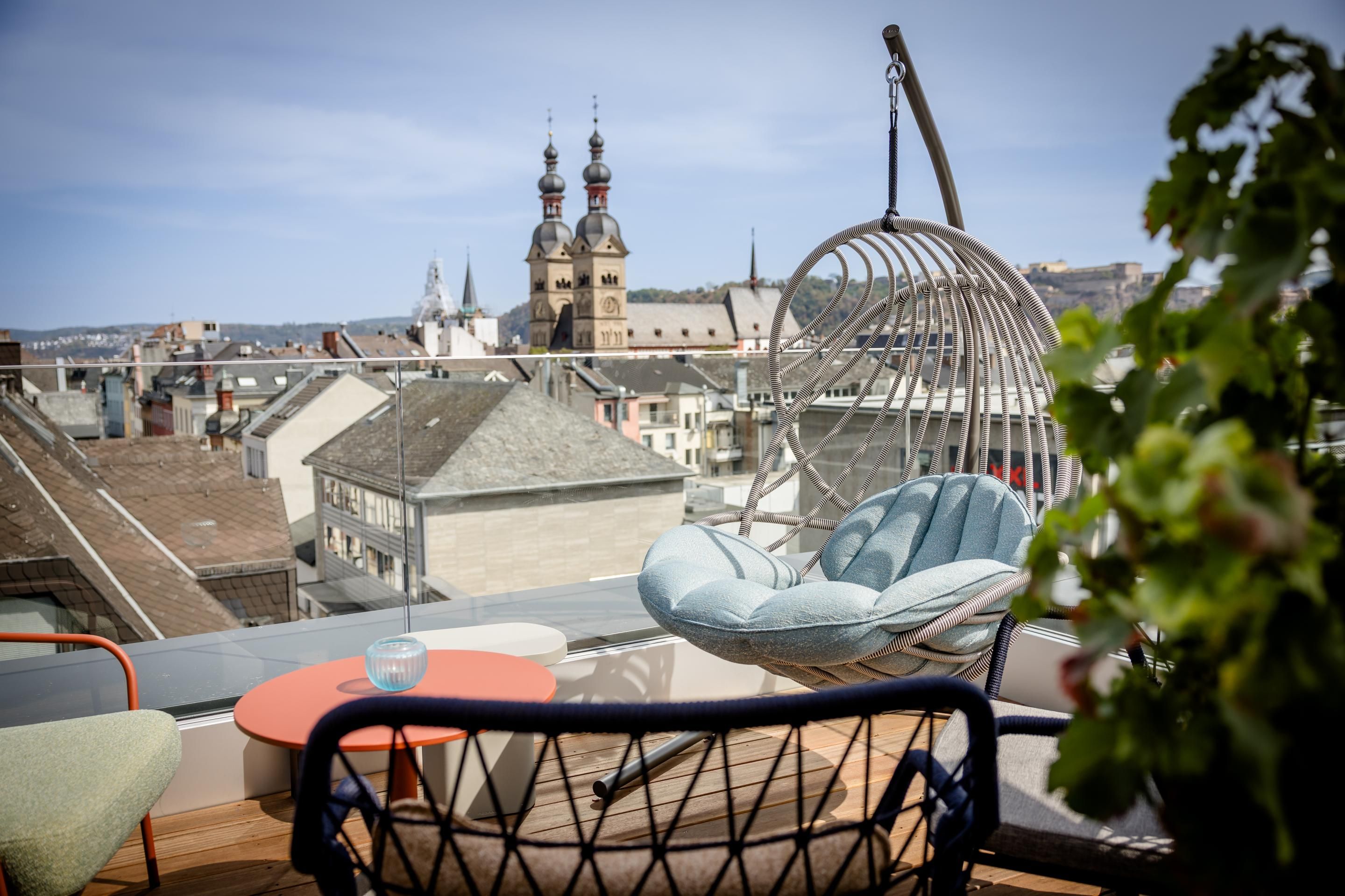 Gemütliche Dachterrasse mit moderner Hängeschaukel und Sitzmöbeln, von der aus sich ein weiter Blick auf historische Stadtdächer und Türme bei klarem Himmel bietet.