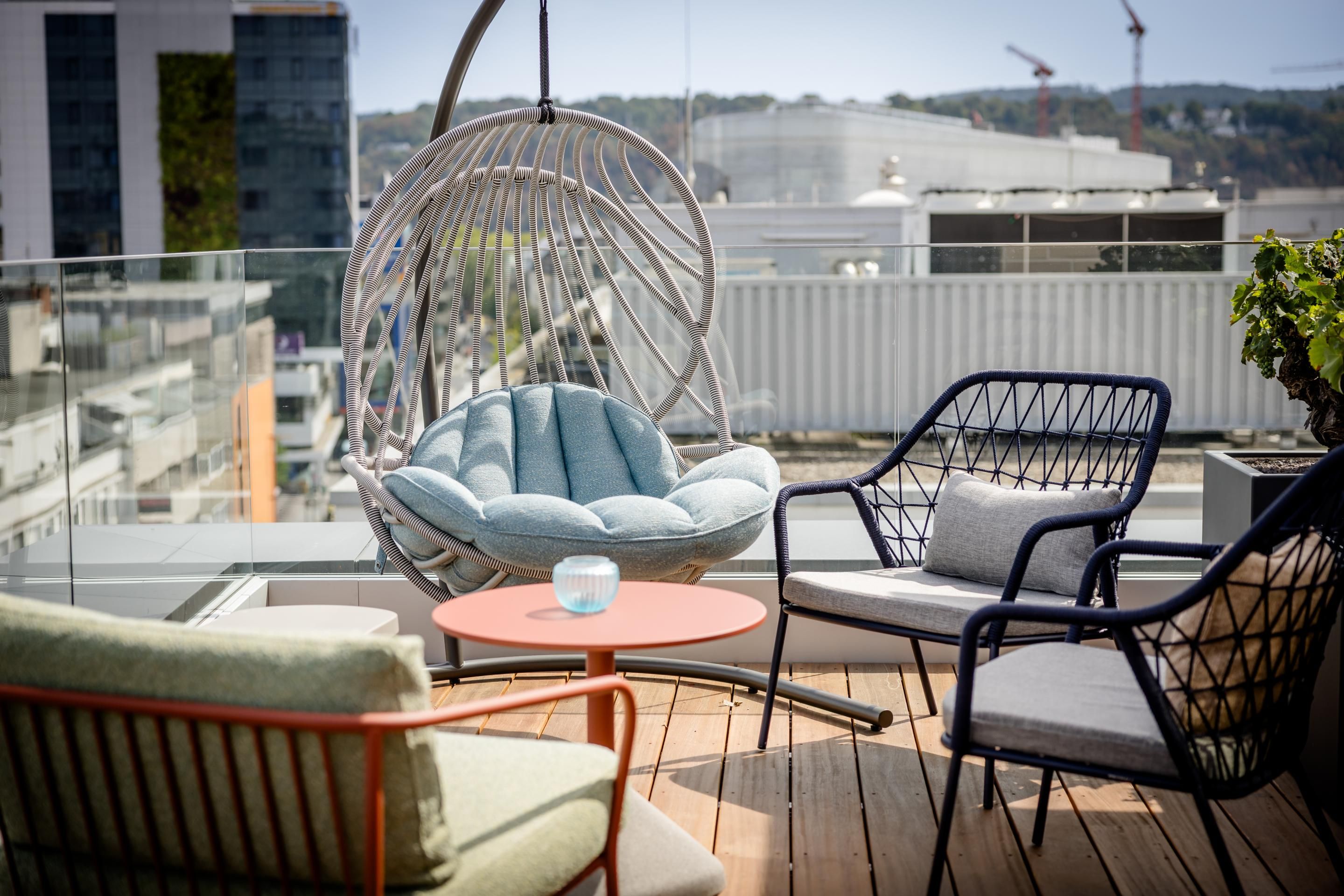 Terrassenbereich mit moderner Sitzgruppe: Hängesessel mit blauem Kissen, zwei schwarze Stühle mit grauen Polstern und ein kleiner runder Tisch, urbaner Ausblick im Hintergrund.
