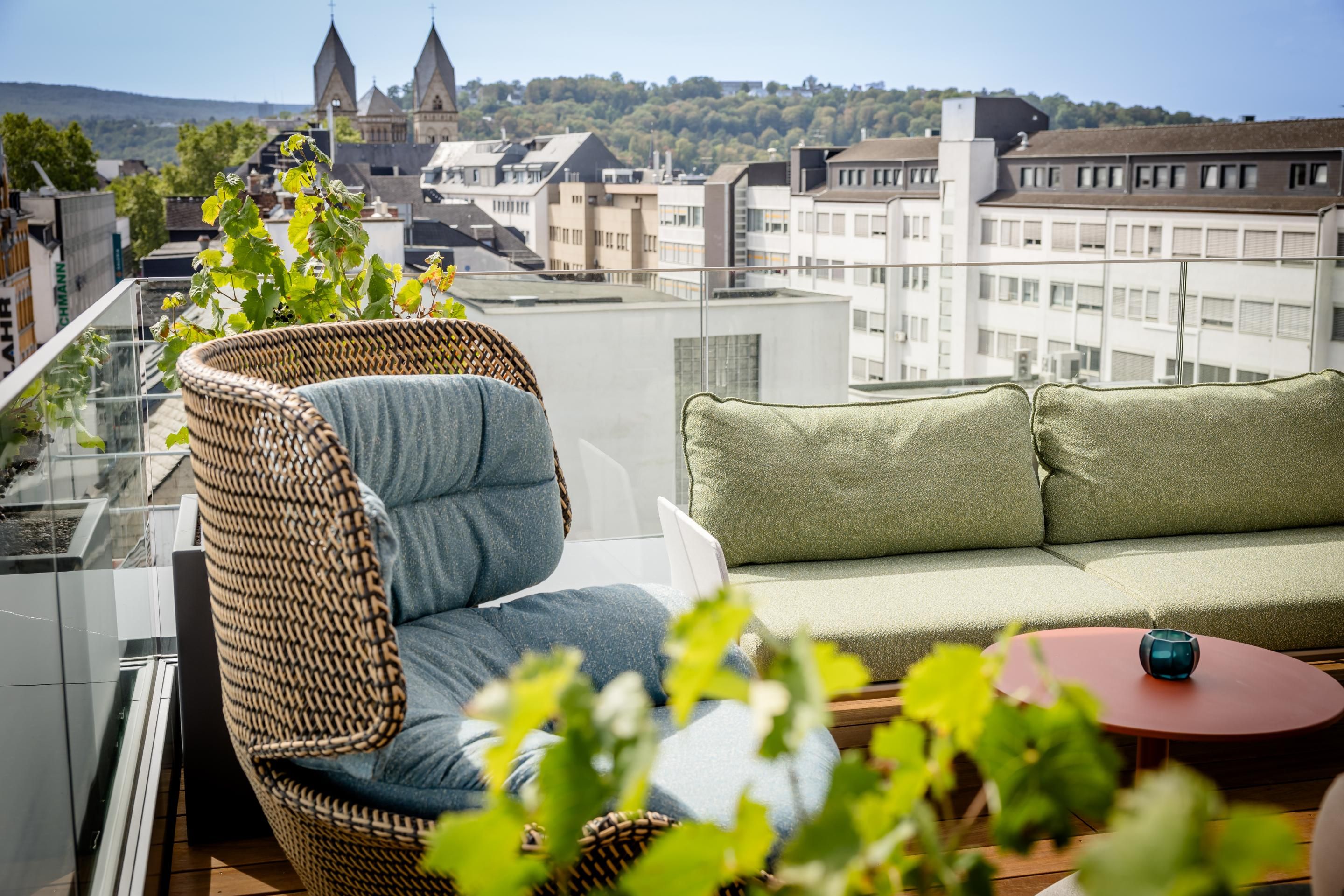 Gemütliche Dachterrasse mit gepolstertem Korbsessel und grünem Sofa, blühenden Pflanzen und Blick auf Stadtgebäude und Kirchtürme am Horizont.