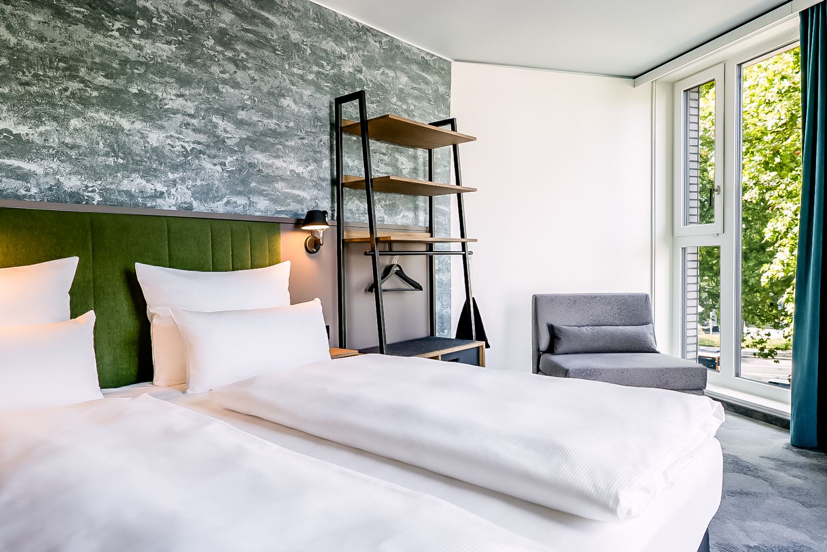 Modernes Hotelzimmer mit zwei Einzelbetten, grünem Kopfteil, offenem Regal und großem Fenster, das viel Tageslicht und Blick ins Grüne bietet.
