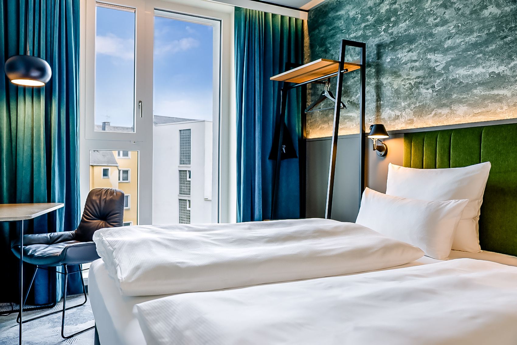 Modern eingerichtetes Hotelzimmer mit großem Fenster, offenem Blick auf Stadtgebäude, gemütlichem Bett mit weißer Bettwäsche, blaugrünen Vorhängen und stilvoller Wandbeleuchtung für entspannte Atmosphäre.