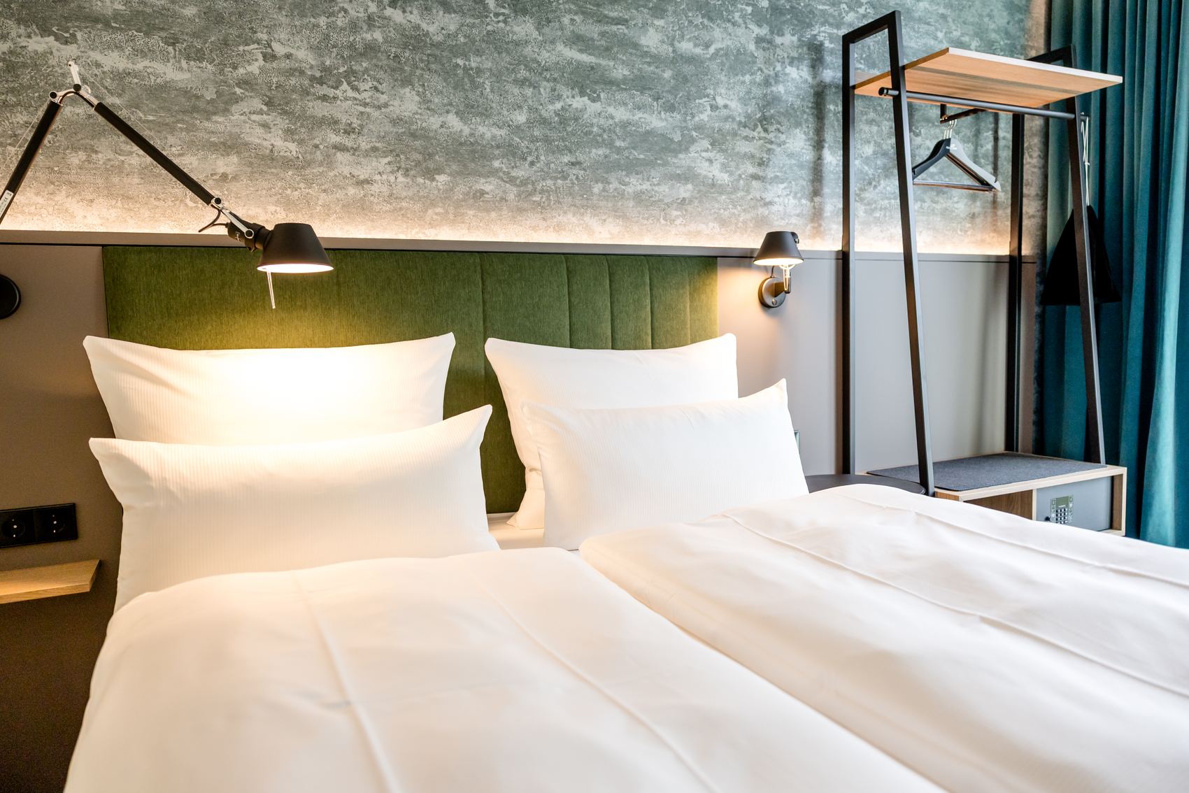 Modern eingerichtetes Hotelzimmer mit zwei Einzelbetten, grüner gepolsterter Kopfteil, warmem Leselicht und offenem Kleiderständer, das eine helle und einladende Atmosphäre vermittelt.