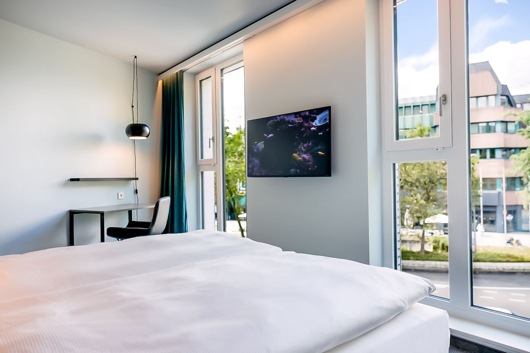 Helles Hotelzimmer mit großem Bett, Schreibtisch und Blick durch bodentiefe Fenster auf die Stadt. Moderne, klare Einrichtung und viel Tageslicht schaffen eine angenehme Atmosphäre für Arbeit und Erholung.
