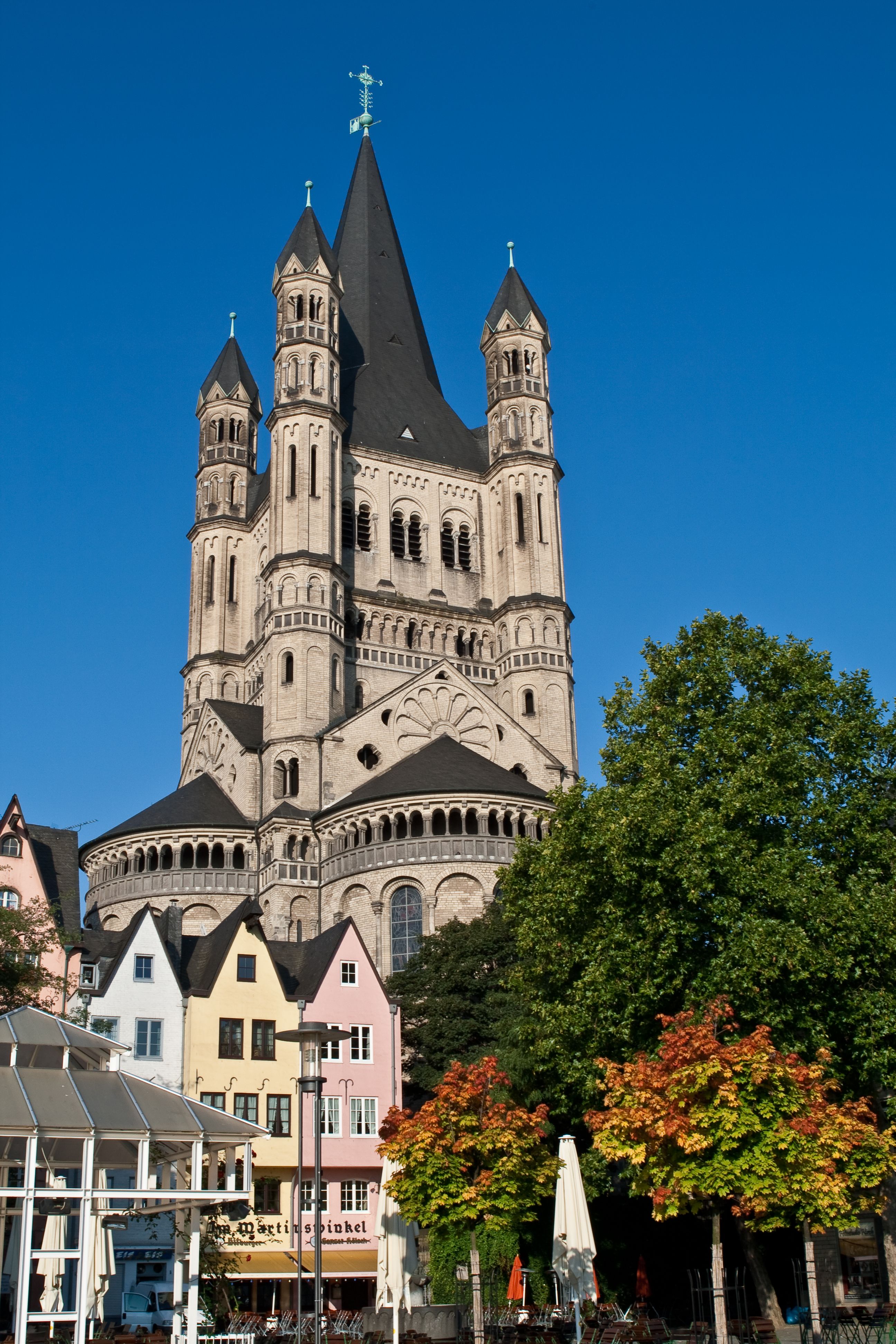 Große romanische Kirche mit hohen Türmen und spitzen Dächern hinter bunten Fachwerkhäusern, umgeben von grünen und herbstlich gefärbten Bäumen, unter klarem blauem Himmel.