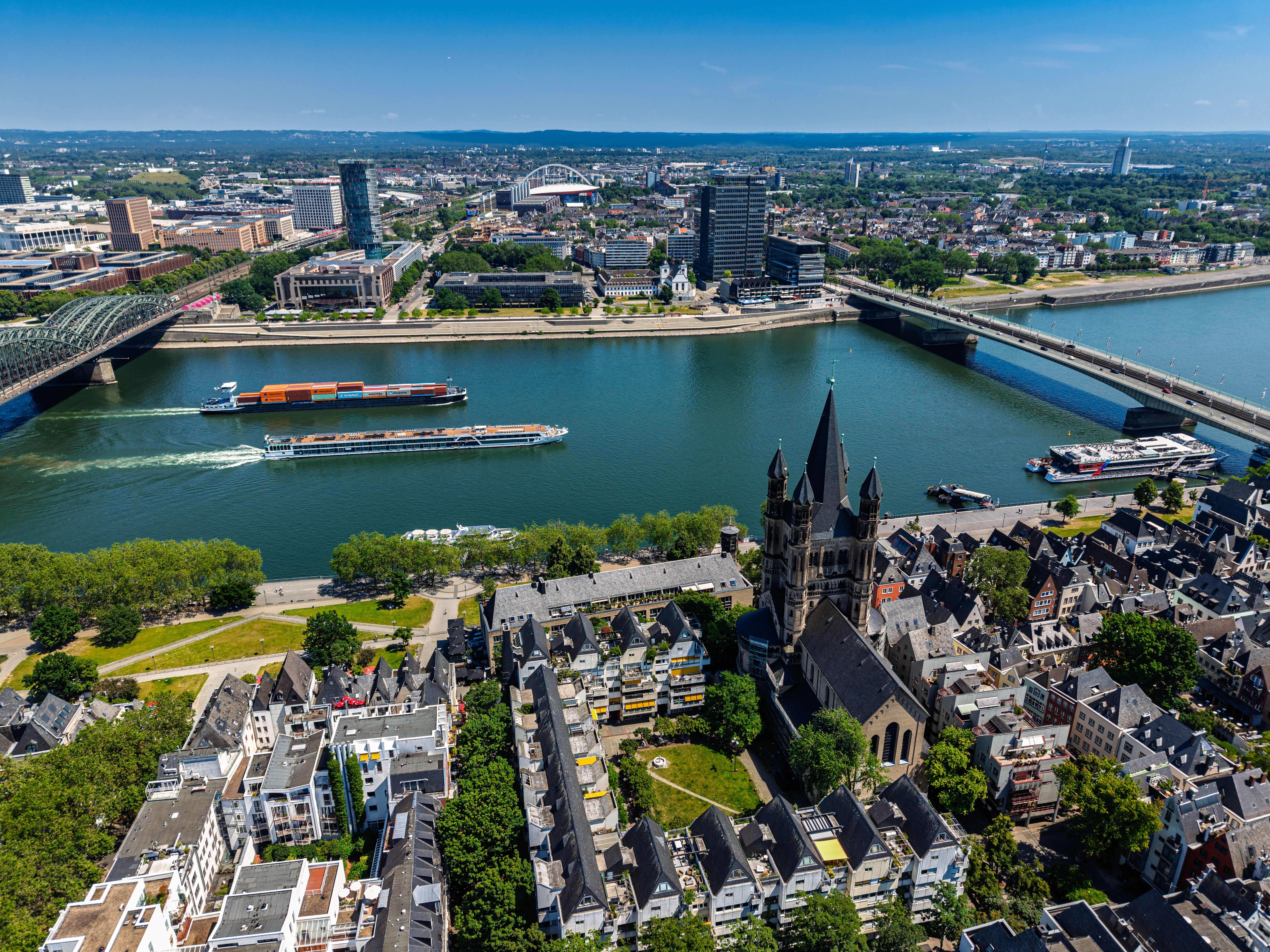 Blick über den Rhein mit vorbeifahrenden Schiffen, zwei Brücken verbinden die Stadtteile, im Vordergrund die historische Altstadt mit einer markanten Kirche und grünen Parkflächen. Klare Sicht über die Stadtlandschaft an einem sonnigen Tag.