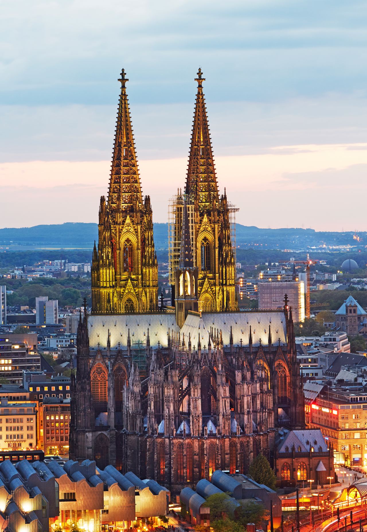 Beeindruckender Blick auf den beleuchteten Kölner Dom bei Abenddämmerung, umgeben von der modernen Stadtlandschaft Kölns.