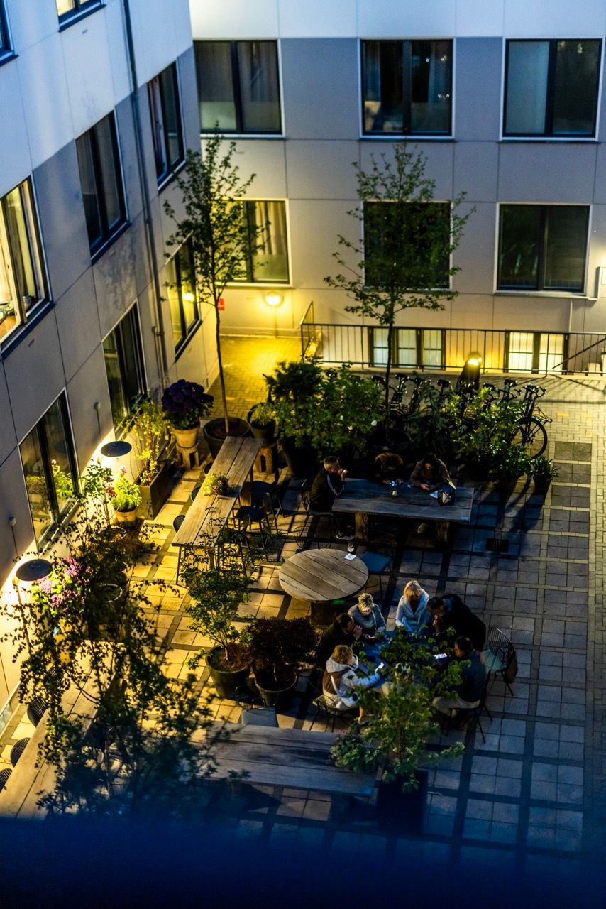 Abendliche Innenhof-Terrasse eines Hotels mit gemütlicher Beleuchtung, mehreren Sitzgruppen und Gästen, die entspannt beisammensitzen, umgeben von Pflanzen und Bäumen.