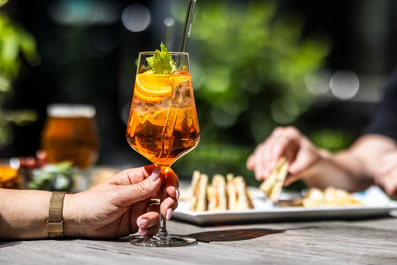 Hand hält ein Glas Aperitif mit Eis, Orangenscheiben und Minzblatt beim sonnigen Außenessen, im Hintergrund Teller mit Snacks und entspannte Atmosphäre.