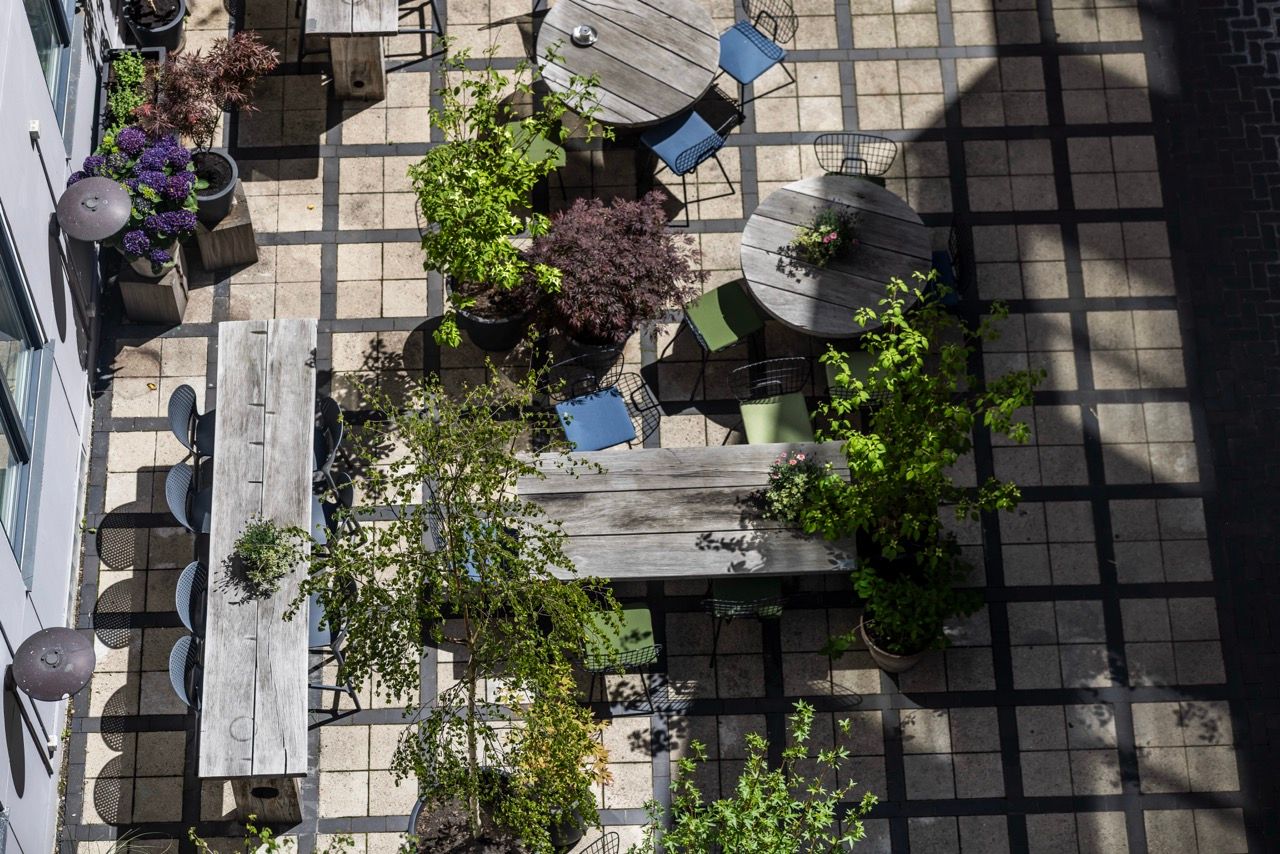 Innenhof mit gepflanzten Bäumen und Sträuchern, umgeben von Tischen und Stühlen aus Holz, mit Sitzgelegenheiten für entspannte Pausen im Freien, bei Sonnenschein.