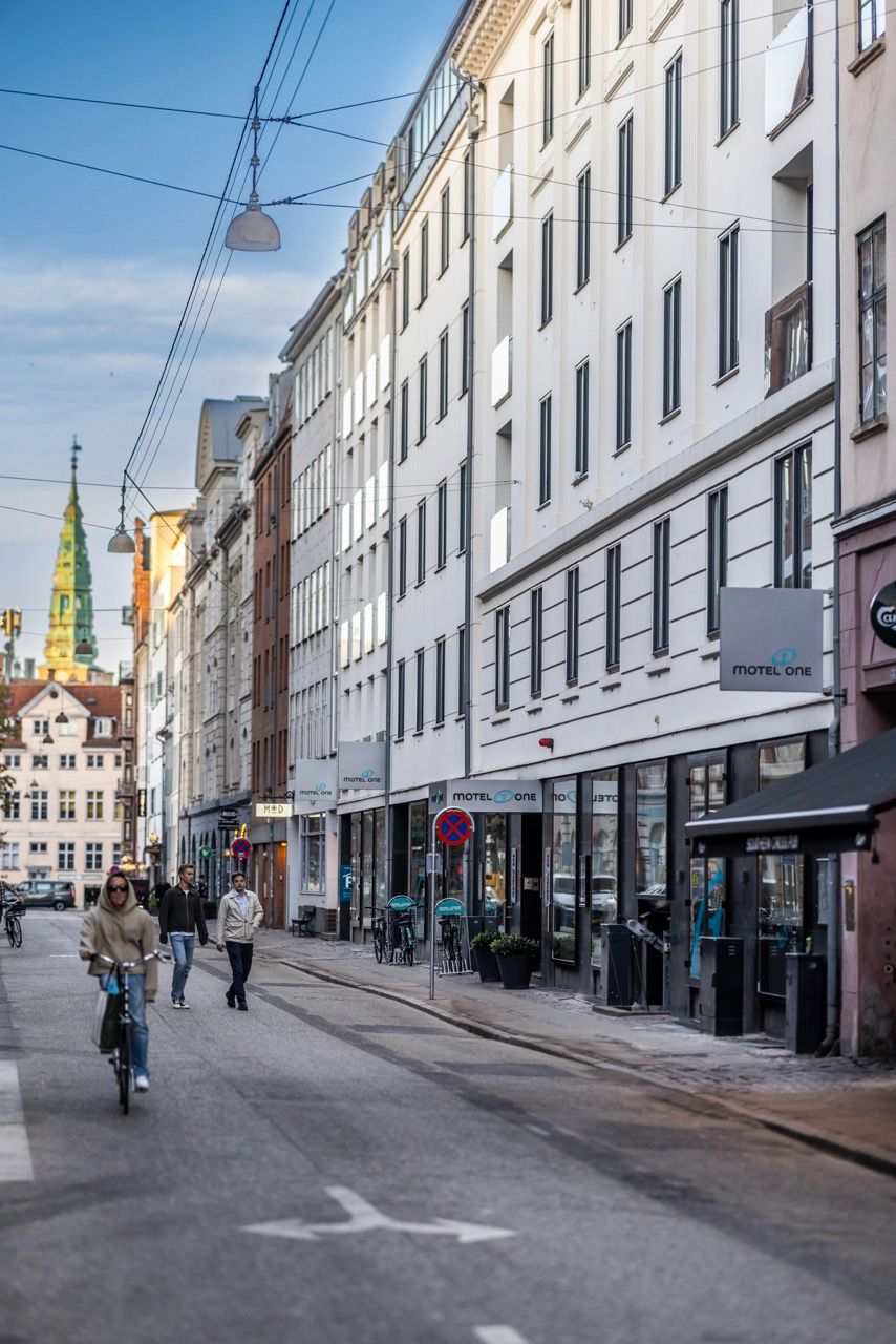 Straßenansicht eines modernen Hotels mit Sitzgelegenheiten im Freien, Passanten und einem Radfahrer, eingebettet in eine belebte städtische Umgebung mit historischen Gebäuden im Hintergrund.