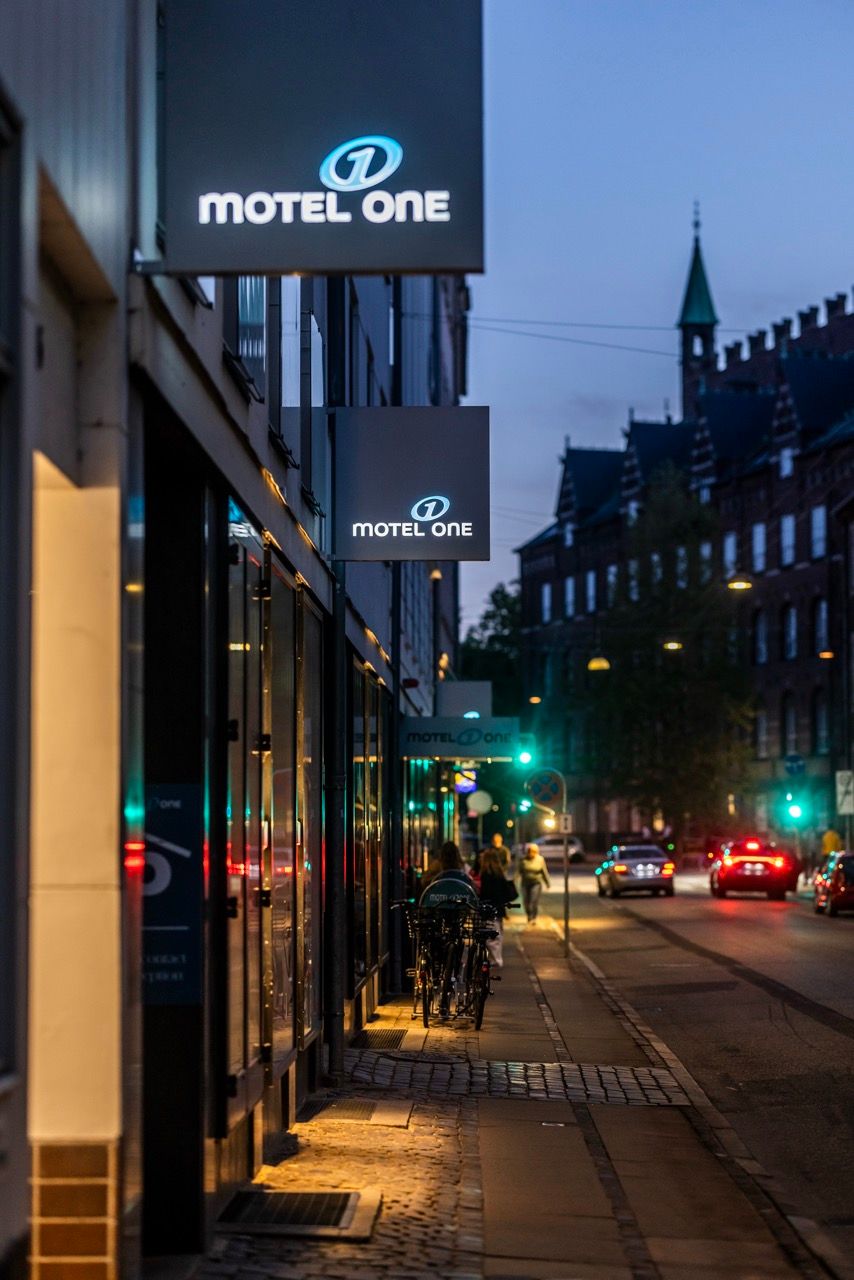 Außenansicht eines Motel One Geschäftsgebäudes am Abend, beleuchtete Hotelschilder, städtische Umgebung mit Gehweg, Fahrrädern und Verkehr.
