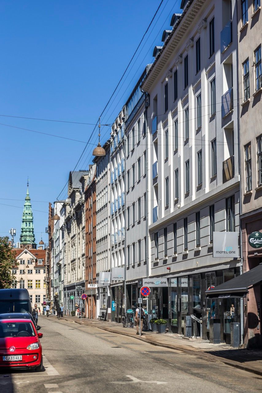 Blick auf eine sonnige Straßenseite mit modernen Hotels und historischen Gebäuden in einer europäischen Stadt, klare blaue Himmel, ruhiger Verkehr, Fußgänger auf dem Gehweg.