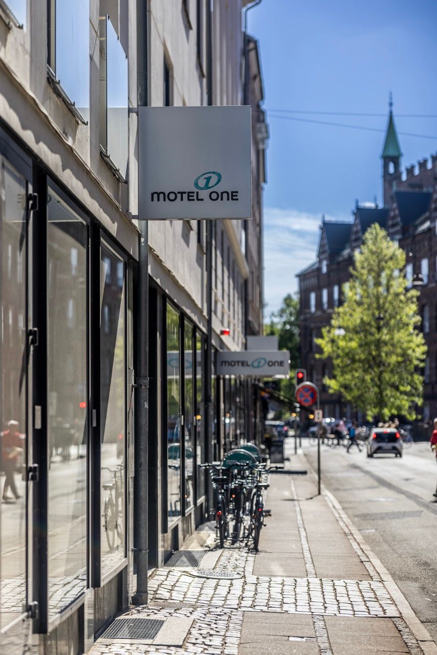 Außenansicht eines Motel One Hotels in einer belebten Stadtstraße, mit Fahrrädern vor dem Eingang und historischen Gebäuden im Hintergrund.