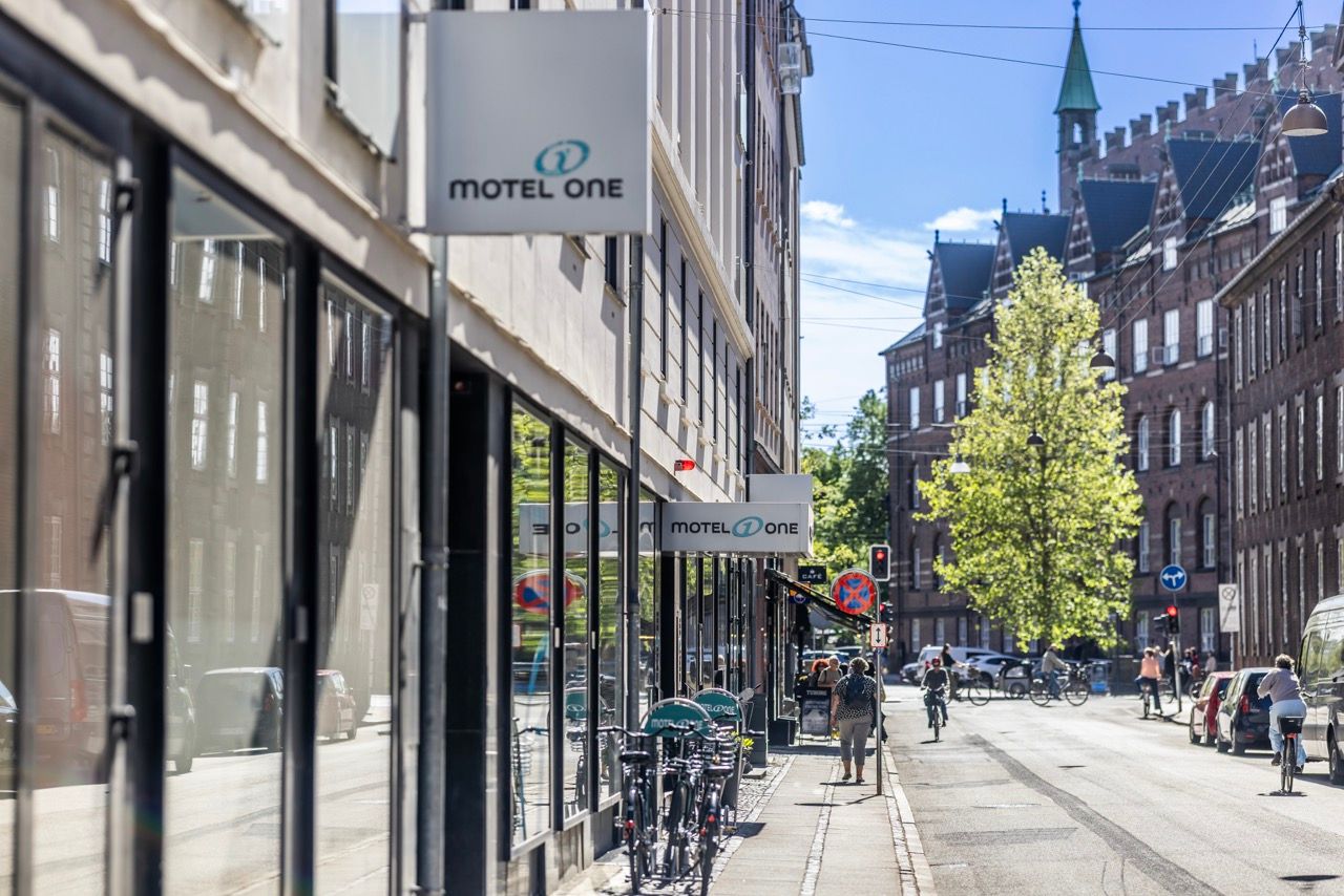 Straße mit dem Motel One Hotelschild an einem modernen Gebäude, Radfahrer und Passanten, sonniger Tag, historische und moderne Architektur kombiniert.