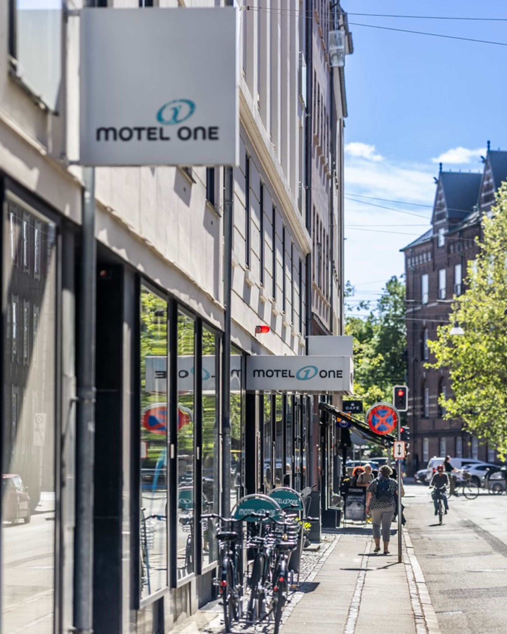 Straße mit dem Motel One Hotelschild an einem modernen Gebäude, Radfahrer und Passanten, sonniger Tag, historische und moderne Architektur kombiniert.