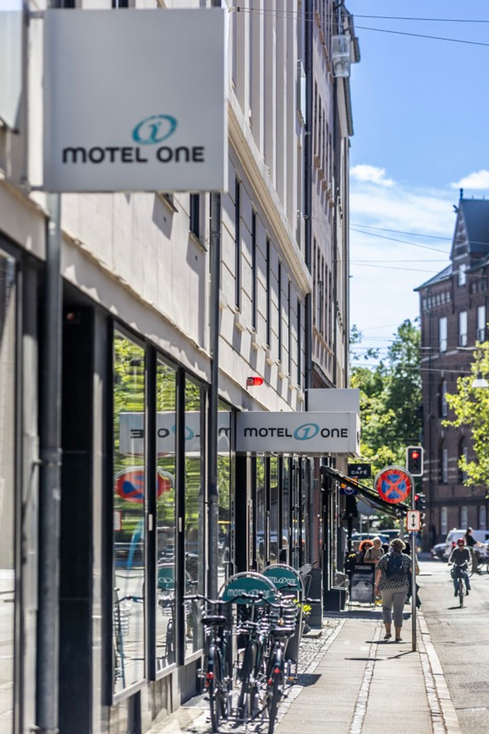 Straße mit dem Motel One Hotelschild an einem modernen Gebäude, Radfahrer und Passanten, sonniger Tag, historische und moderne Architektur kombiniert.
