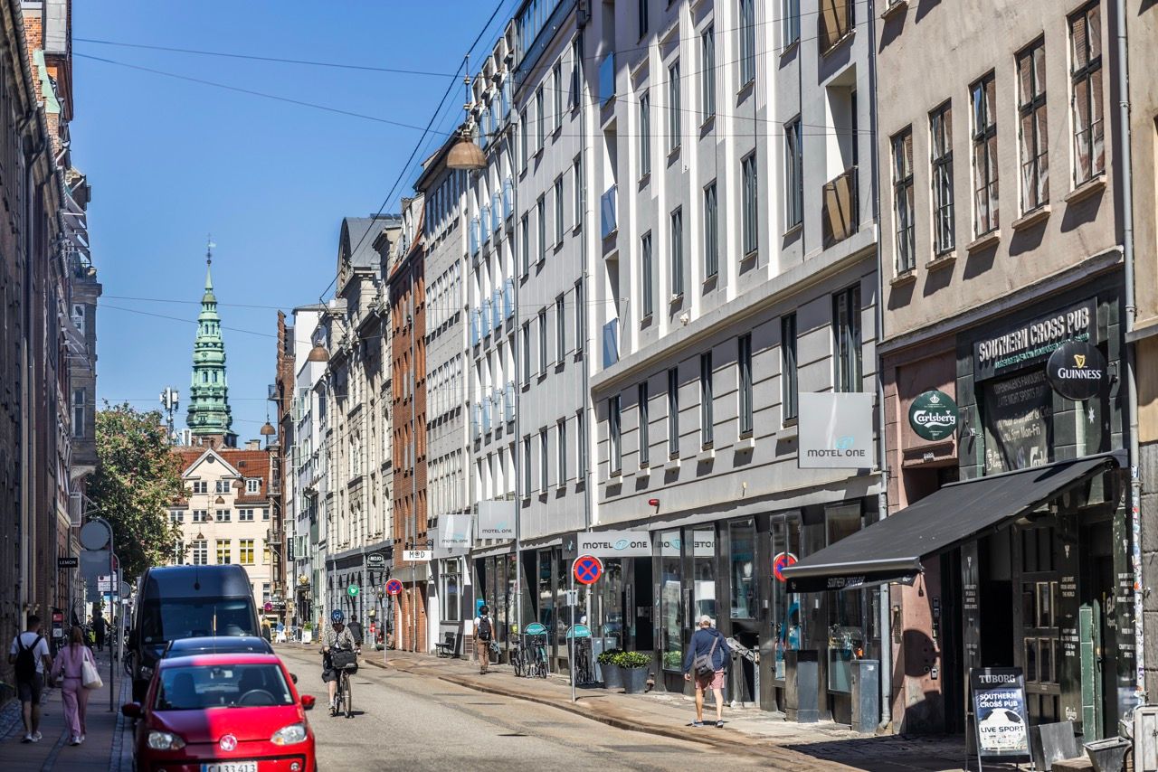Blick auf eine belebte Stadtstraße mit historischen und modernen Gebäuden, Fußgängern und Fahrzeugen, im Hintergrund ein markanter Turm unter blauem Himmel.