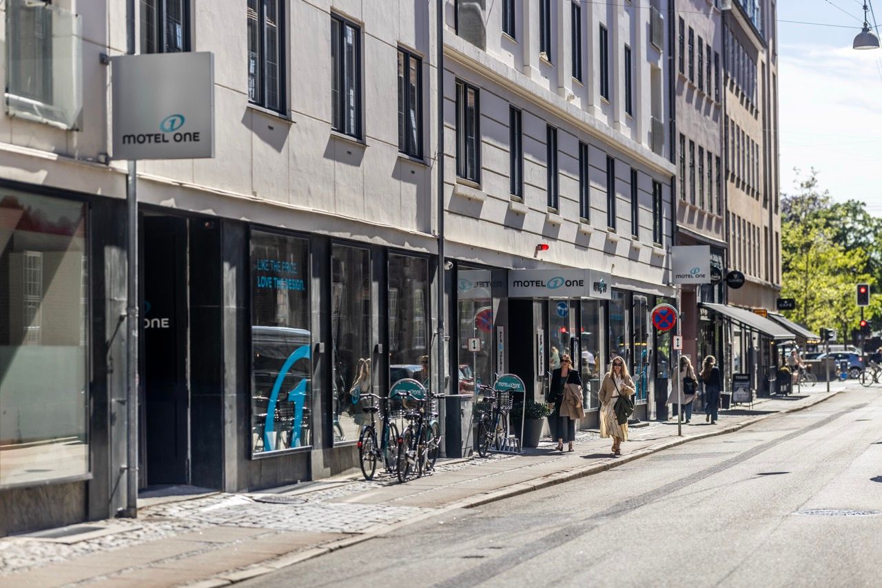 Straßenszene mit Eingang und Beschilderung des modernen Motel One Hotels in einem urbanen Viertel, Passanten gehen auf dem Bürgersteig entlang.