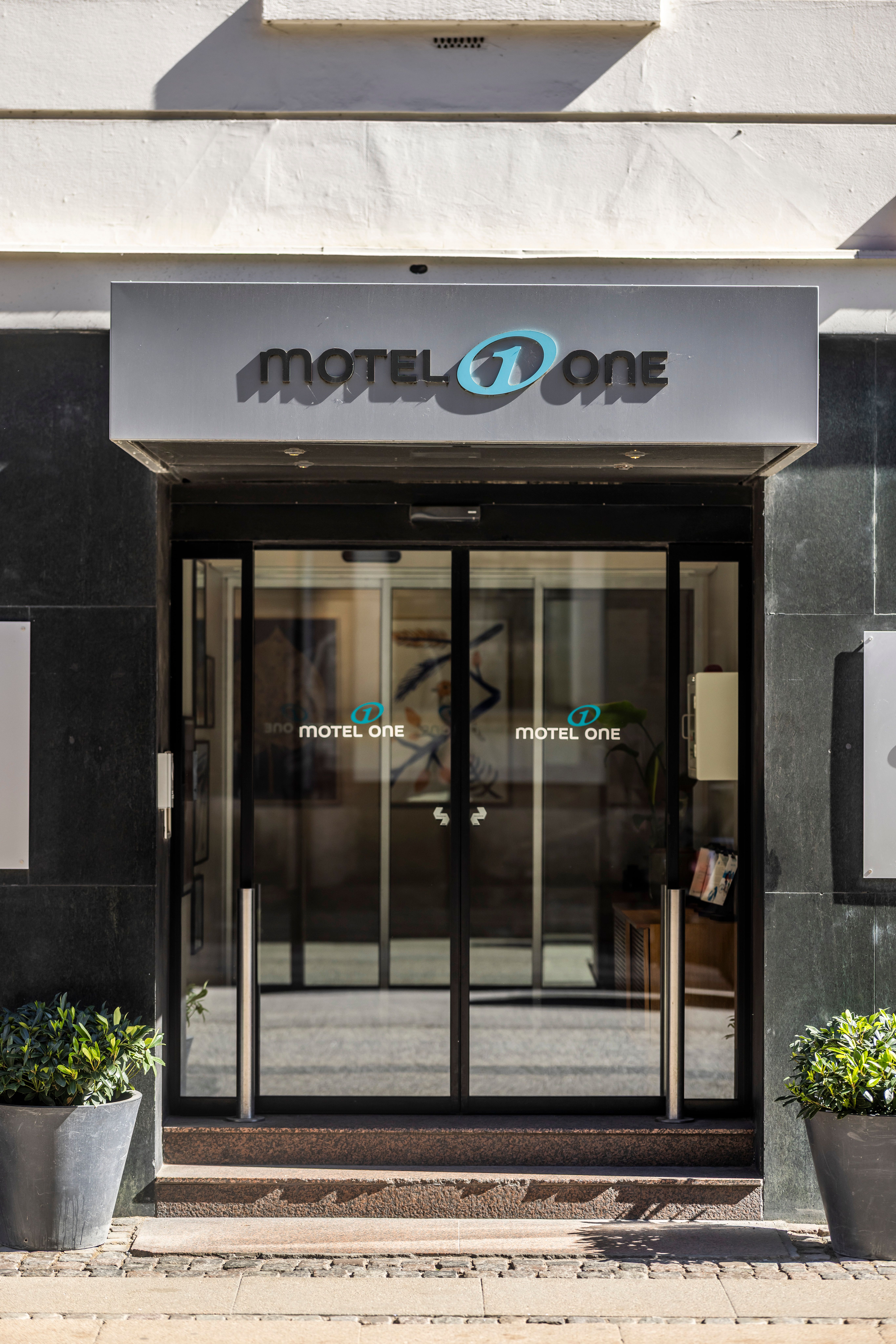 Eingang eines Motel One Hotels mit gläserner automatischer Schiebetür und sichtbarem Hotel-Logo über dem Eingang, flankiert von Pflanzen in großen Töpfen.