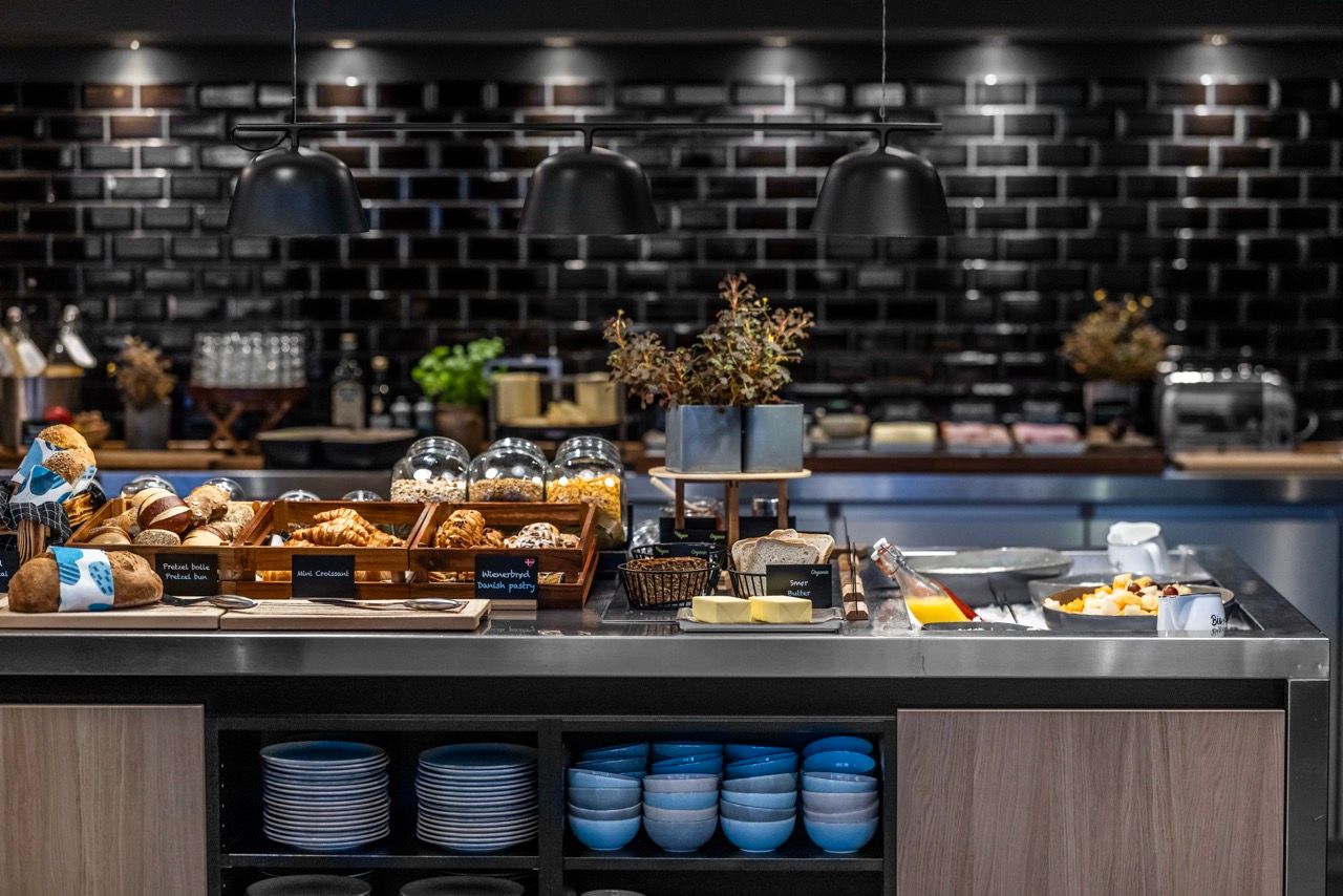 Buffet mit einer Auswahl frischer Backwaren, Aufschnitt, Käse und Butter, stilvoll präsentiert auf einer modernen Theke vor einer schwarzen Ziegelwand, mit warmem Licht für ein einladendes Frühstückserlebnis.