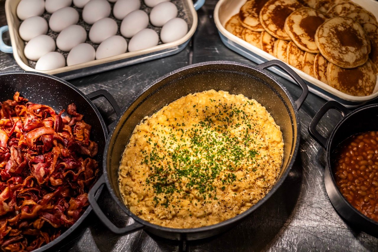 Verschiedene Frühstücksoptionen: Rührei mit Kräutern, knuspriger Frühstücksspeck, weiße Eier, Pancakes und baked beans, serviert in warmen Schalen.