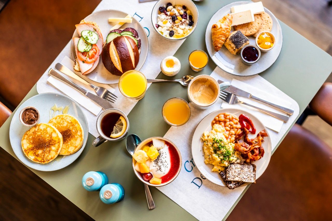 Frühstückstisch mit abwechslungsreichem Angebot: Pancakes, Rührei mit Speck, Brötchen mit Lachs, Müsli, frisches Obst, Croissants, Käse, Marmelade, Kaffee und Orangensaft, liebevoll angerichtet für einen genussvollen Start in den Tag.